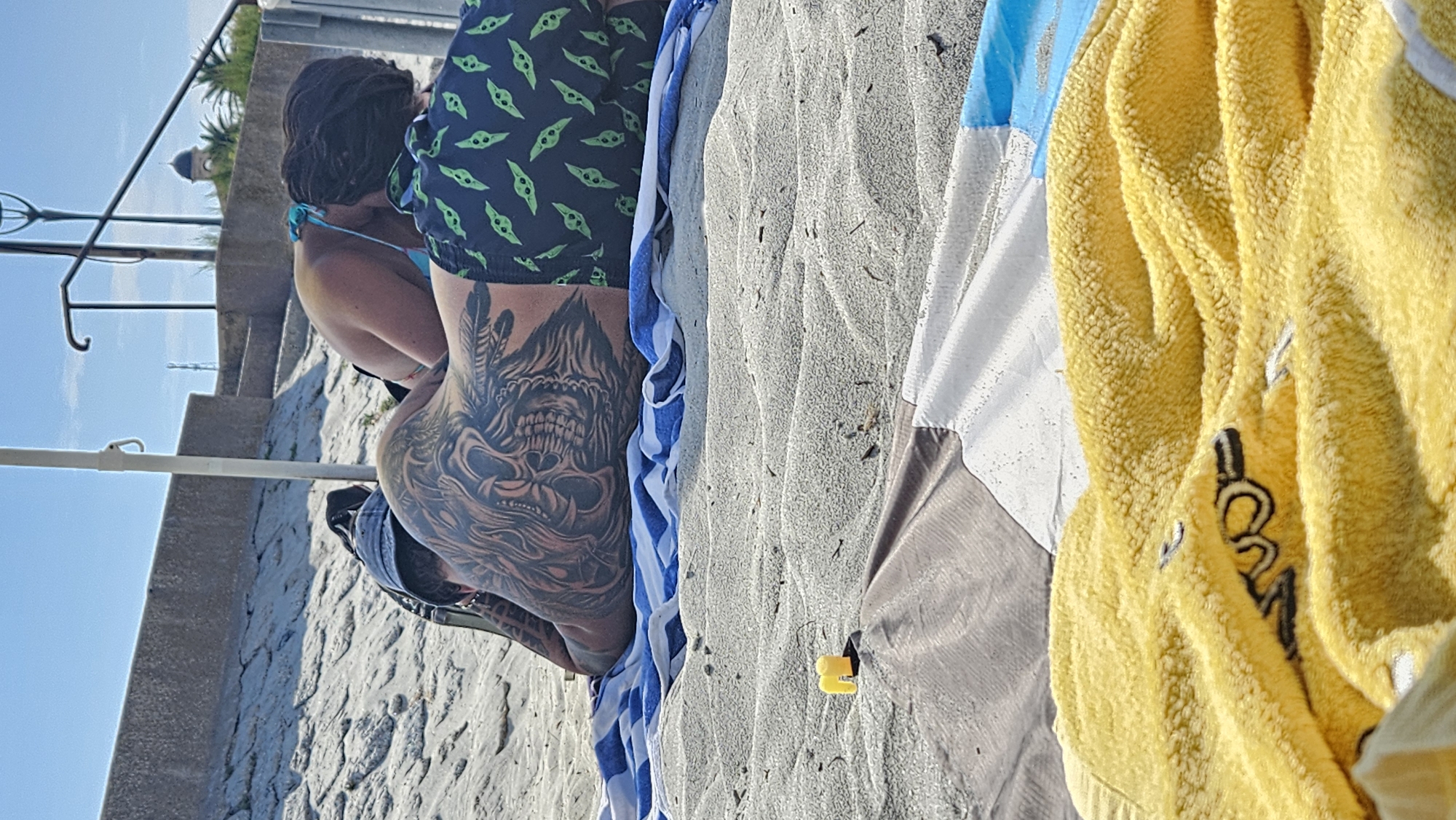 The image depicts a beach scene in L'Île-Rousse, France, under a clear blue sky. In the foreground, a yellow towel with a black design is draped over a beach umbrella or mat. Behind this, a blue and white striped towel lies on the sand. Two people are visible, lying on the beach. The person in the foreground is male, seen from the back, with extensive tattoos covering their back and arms. They are wearing dark swim trunks with a repeating pattern of green figures. Next to them, a person identified as female is seen from the back and side, wearing a blue bikini top. They are leaning forward, their face obscured, possibly interacting with the male. The background shows a textured concrete wall, a set of stone steps, and a distant lamppost. The sand is light in color and appears rippled.