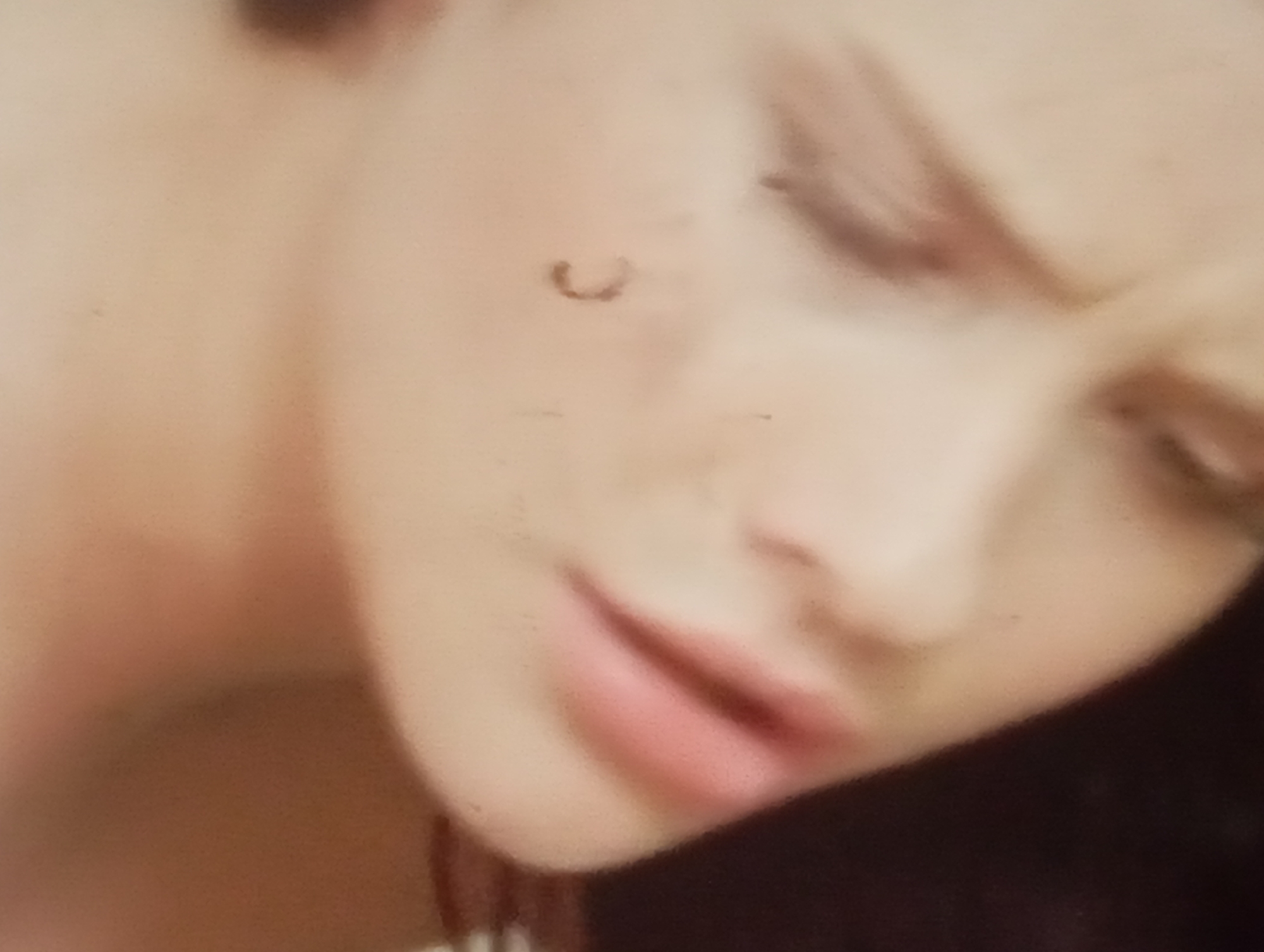 A close-up, blurry photograph shows the left side of a person's face, including their cheek, eye, and mouth. The person has light skin and appears to be wearing makeup on their lips. There is a small, dark mark or blemish on the cheek. The background is out of focus and dark, suggesting an indoor setting. The image was taken in Sofiivska Borshchahivka, Ukraine.