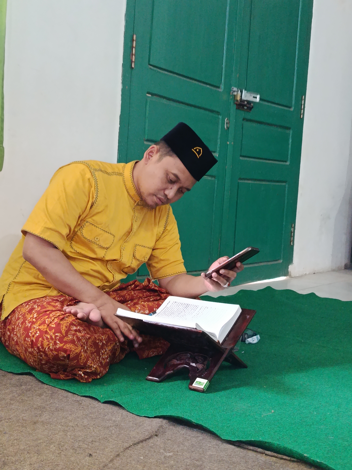 A man wearing a yellow shirt and a black cap with a gold emblem is sitting on a green rug, reading a book placed on a wooden stand. He is holding a mobile phone in his right hand. The man is positioned in front of a green double door. The environment appears to be indoors, possibly in Bondowoso, Indonesia, suggested by the distinct architectural elements like the wooden door and the flooring. The man's attire and the presence of the book suggest a religious or scholarly activity.