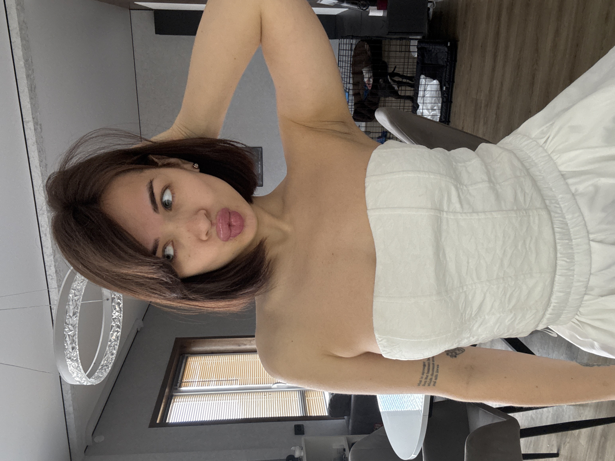 A woman with brown hair and fair skin poses indoors, wearing a white, strapless top. She has her right arm raised, her hand near her hair, and is making a puckered lip expression. Her left arm is down, with a visible tattoo. A circular chandelier is visible in the background, along with a window with blinds, chairs, and a pet crate. The location is potentially Лобаново, Russia.
