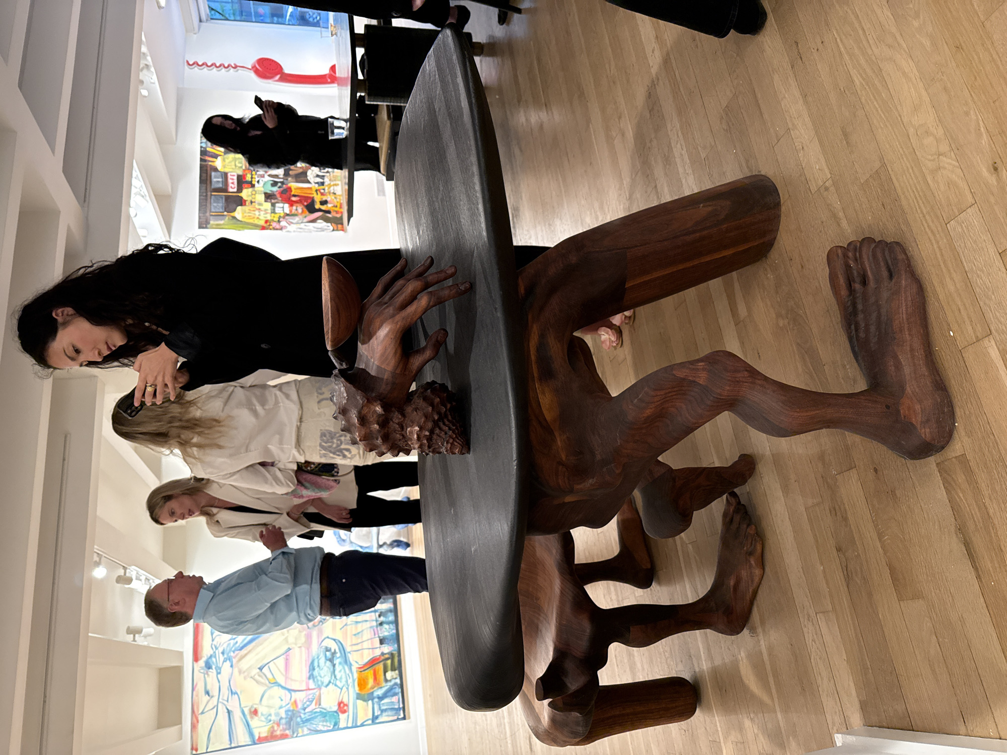 The image captures a scene inside an art gallery in New York City, United States of America. The predominant feature is a large, dark wooden sculpture resembling human figures supporting a wide, oval-shaped top, possibly serving as a table. Details of the sculpture include intricately carved hands, feet, and limbs.
Several people are observing the sculpture. A woman with dark hair is closely examining it, while two women wearing white jackets and a man in a light blue shirt and dark pants are looking at other artwork in the gallery. The environment includes light-colored wooden flooring, white walls, and other paintings displayed on the walls. A red telephone sculpture is visible in the background, along with additional artwork. The lighting is bright, suggesting a well-lit gallery space.
