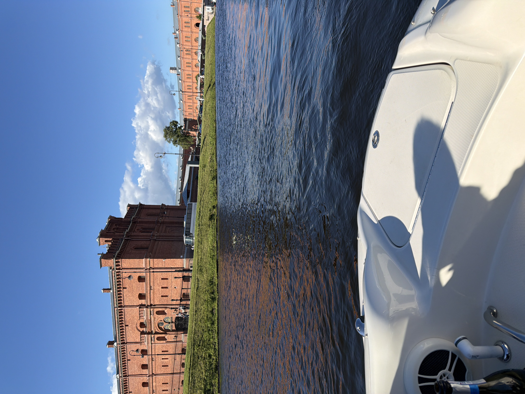 The image shows a view from a boat on a body of water looking towards a large, red brick building. The building has a distinctive architectural style with multiple arched windows and what appear to be turrets or decorative elements on the roofline. The building is set against a backdrop of a bright blue sky with scattered white clouds. To the right of the main building, a longer, also red brick structure is visible, extending along the waterfront. A grassy embankment separates the building from the water. In the foreground, the white interior of a boat is visible, including a speaker and part of a bottle. The water is dark and rippled, reflecting the sunlight and the colors of the sky and buildings. A few people are standing near the entrance of the main building. A white bus is parked on the road behind the grassy embankment.