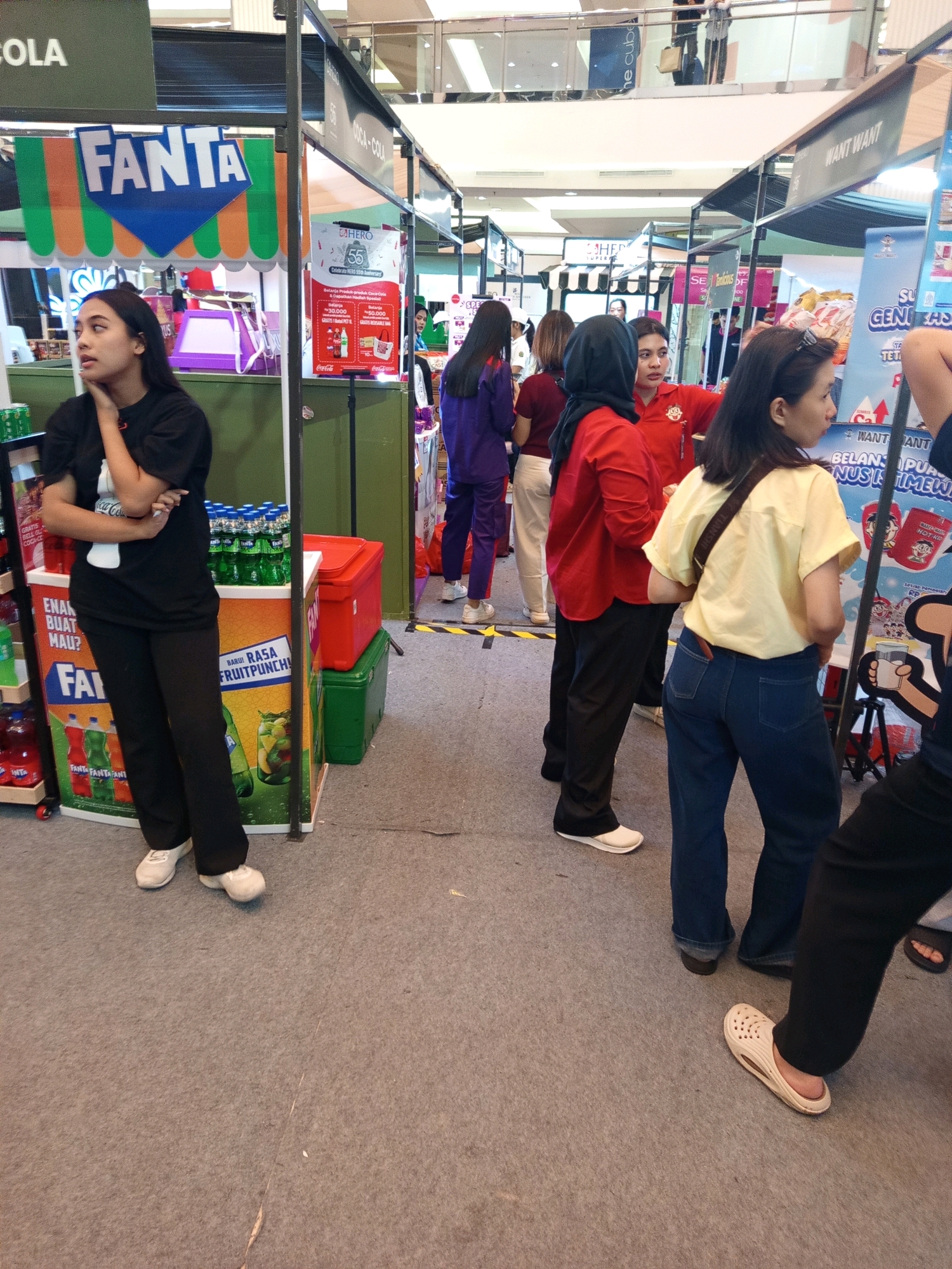 The image depicts an indoor market or exhibition hall in West Jakarta, Indonesia. Several individuals are present, some appearing to be staff at various booths. In the foreground, a young woman with long black hair is standing, wearing a black t-shirt and black pants, leaning against a Fanta branded display. Behind her, other booths are visible, one promoting Coca-Cola and another with a banner for "Want Want". People are seen browsing or working at these stalls. The environment is a commercial space with carpeting on the floor and various product displays. The overall atmosphere suggests a promotional event or a shopping area.