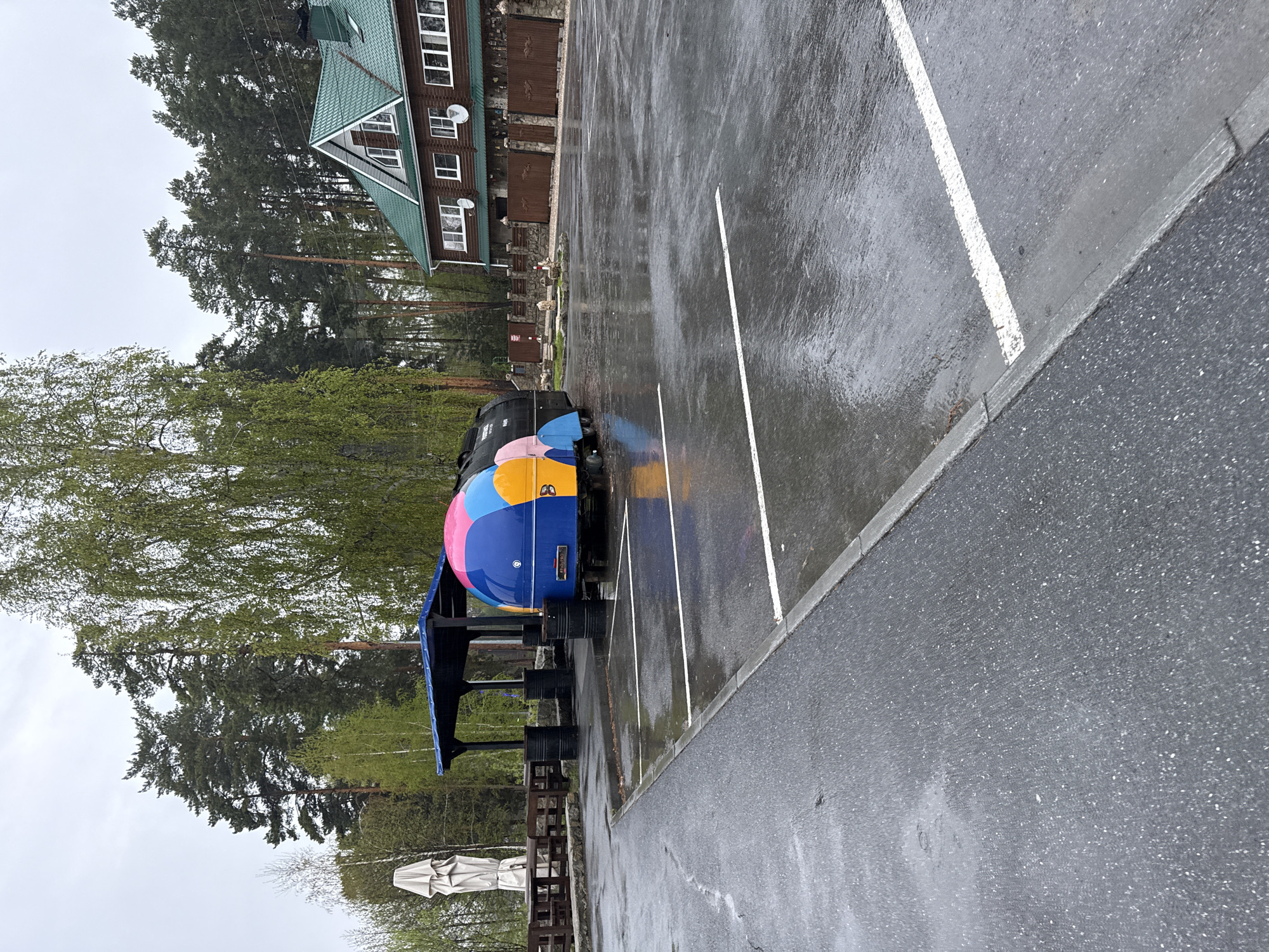 On a rainy day in Turgoyak, Russia, a colorful, round food truck sits parked in a paved parking lot. The truck is painted with bright blue, yellow, and pink colors. Behind the truck, there's an outdoor seating area with tables and umbrellas. In the background, a wooden building with a green roof is visible, surrounded by trees. The parking lot has white painted lines marking parking spaces. The pavement is wet from the rain, reflecting light.