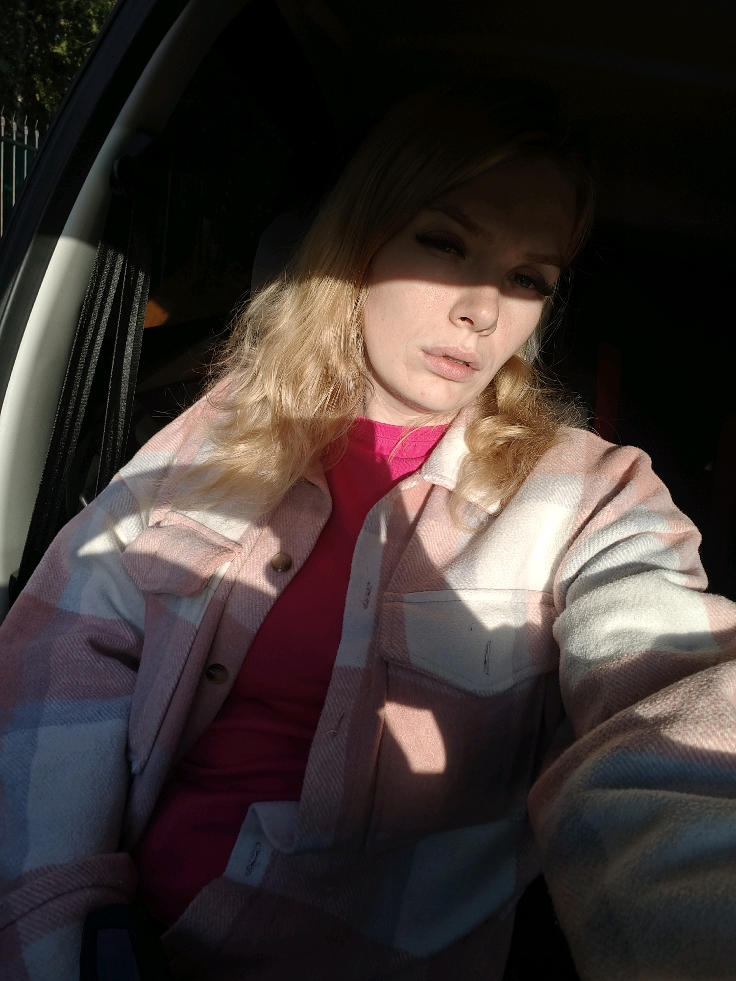 A young woman with blonde hair and light skin sits in the driver's seat of a car, looking at the camera. She is wearing a pink t-shirt under a pink and white plaid shirt. Sunlight streams through the car window, casting shadows on her face and upper body. The interior of the car is dark, with a black seatbelt visible. Outside the car, a green metal fence and trees can be seen. The car appears to be parked on a street in Izhevsk, Russia.