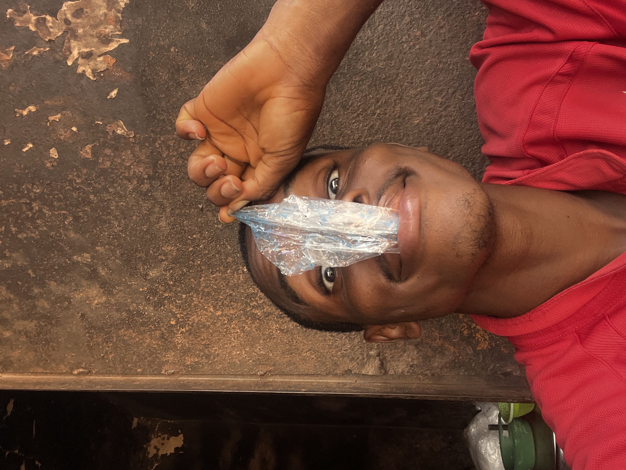 A man lies on a surface in an unknown location in Onitsha, Nigeria. A clear plastic bag covers his nose and mouth, held in place by a hand. He wears a red shirt. The surface appears to be metal with discoloration. Below the surface, partial objects are visible.