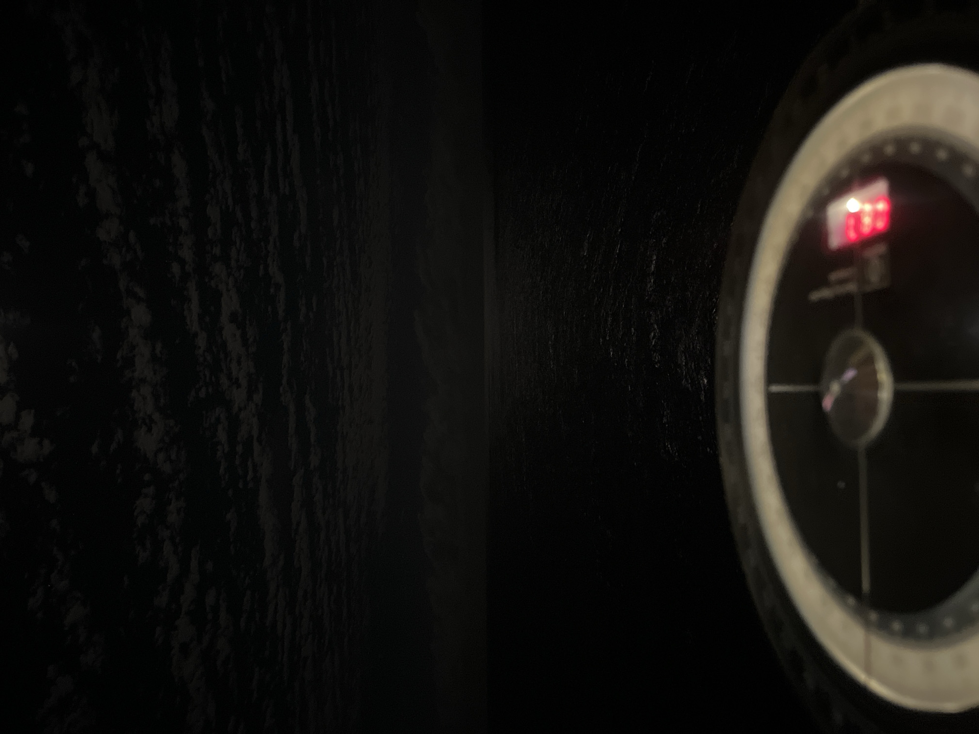 The image depicts a dark, seemingly confined space. On the right, there's a blurry instrument panel with a circular gauge, likely a navigational or depth indicator, displaying a red digital reading. The left side of the image is dominated by a dark, textured surface, possibly a wall or rock formation. The overall environment suggests a submarine or underwater vehicle exploring a dark, undefined location. The city is unknown and the location is unknown.