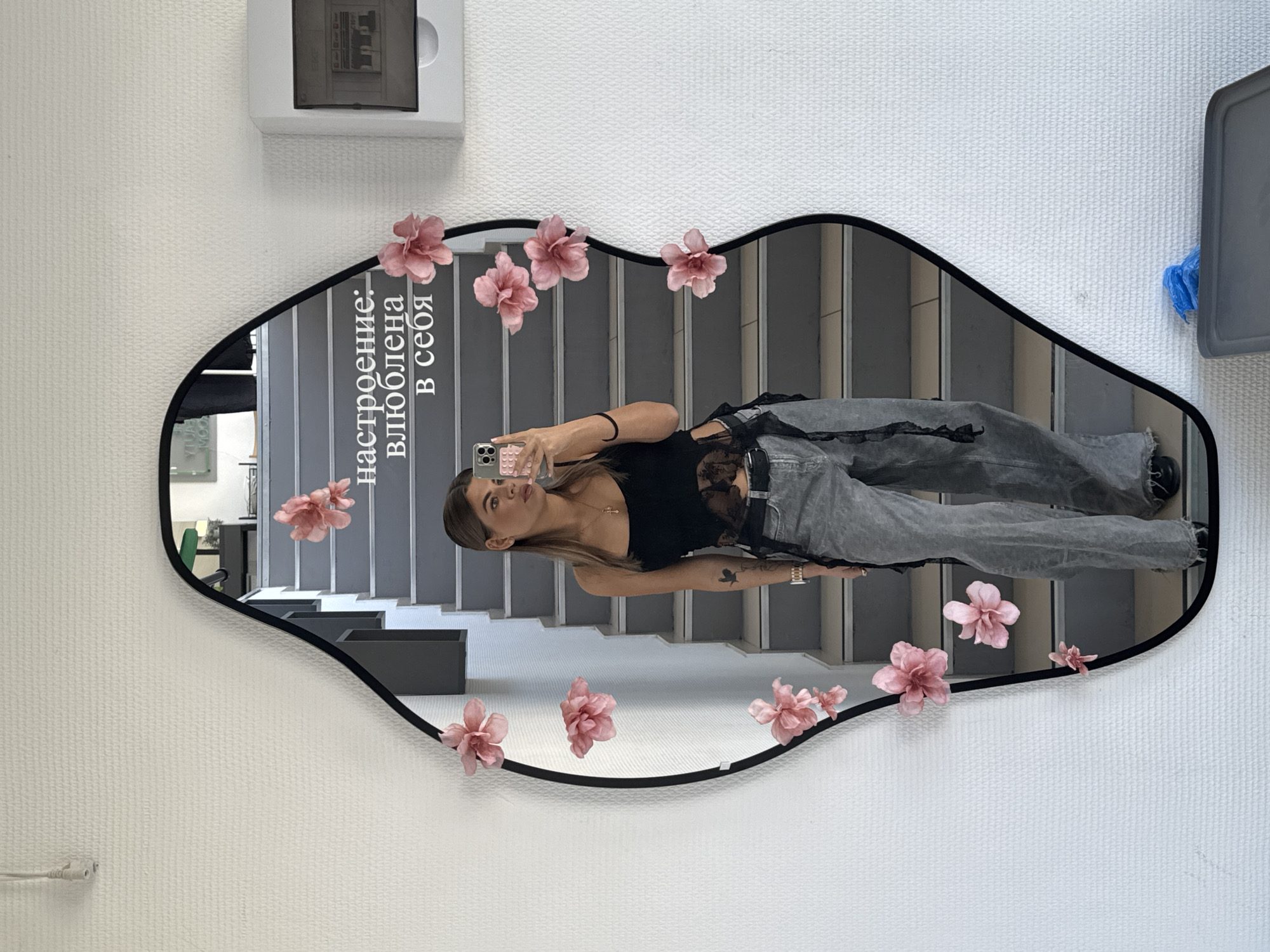 A young woman with light brown hair and fair skin is taking a selfie in a decorative mirror. The mirror has a black frame and is adorned with pink flowers. The reflection shows a stairwell with gray steps and a white wall behind it. The woman is wearing a black top and gray jeans. The inscription on the mirror reads "настроение: влюблена в себя." A white box is mounted on the wall above the mirror. Another container is to the right of the mirror. It is likely located in Chaikovsky, Russia, given the inscription on the mirror.