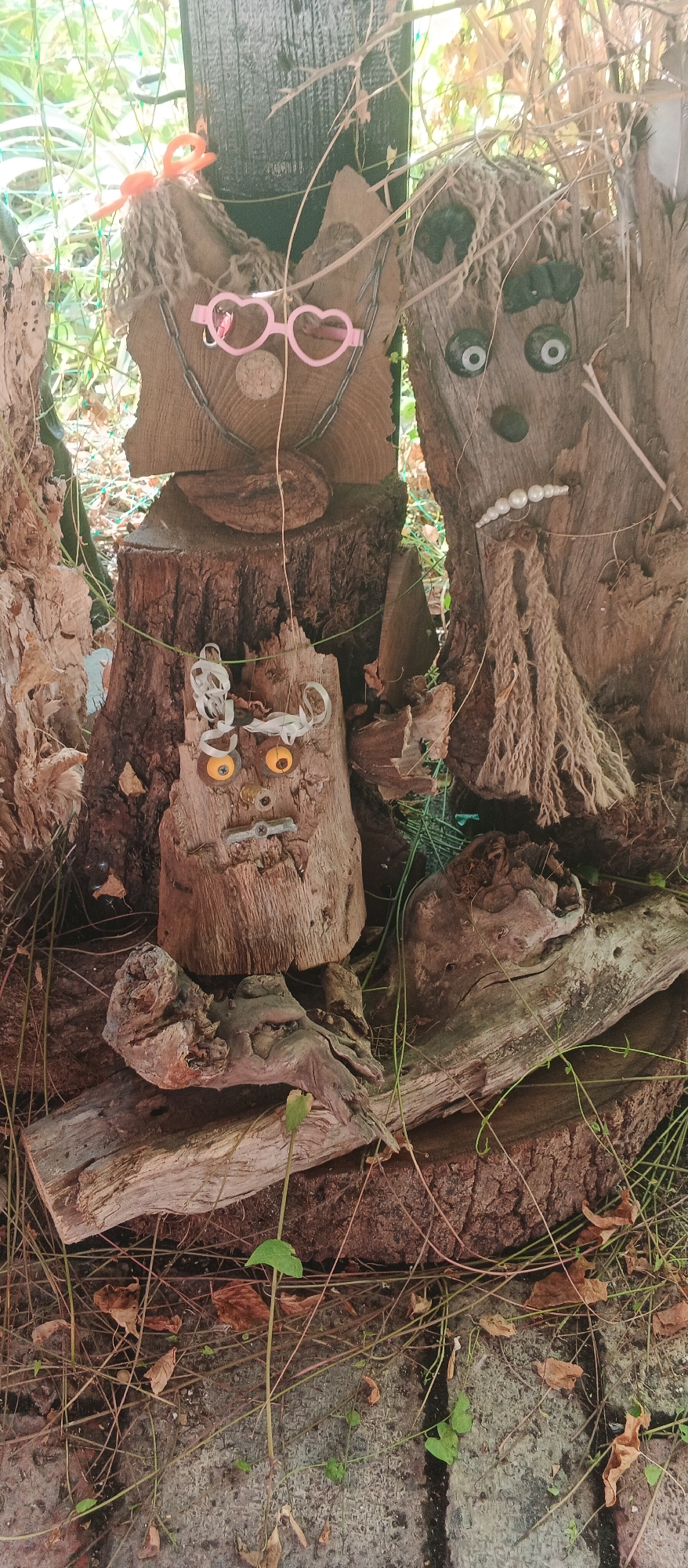 The image displays an outdoor scene in Berehove, Ukraine, featuring a collection of handcrafted figures made from wood and various found objects, arranged among logs and foliage. Two prominent figures, resembling faces, are constructed from thick pieces of wood. The figure on the left has pink heart-shaped glasses, yarn hair, and a chain around its neck. The figure on the right has dark circles for eyes, small black shapes for ears, and a row of white beads for a mouth, giving it a somewhat sad expression. Below these, a smaller wooden figure is assembled with yellow bottle caps for eyes, a small screw for a nose, and a piece of twisted metal for a mouth. The figures are set against a backdrop of rough-hewn logs and branches, with green vines and dry leaves scattered on the ground. The overall impression is of a whimsical, rustic art installation.
