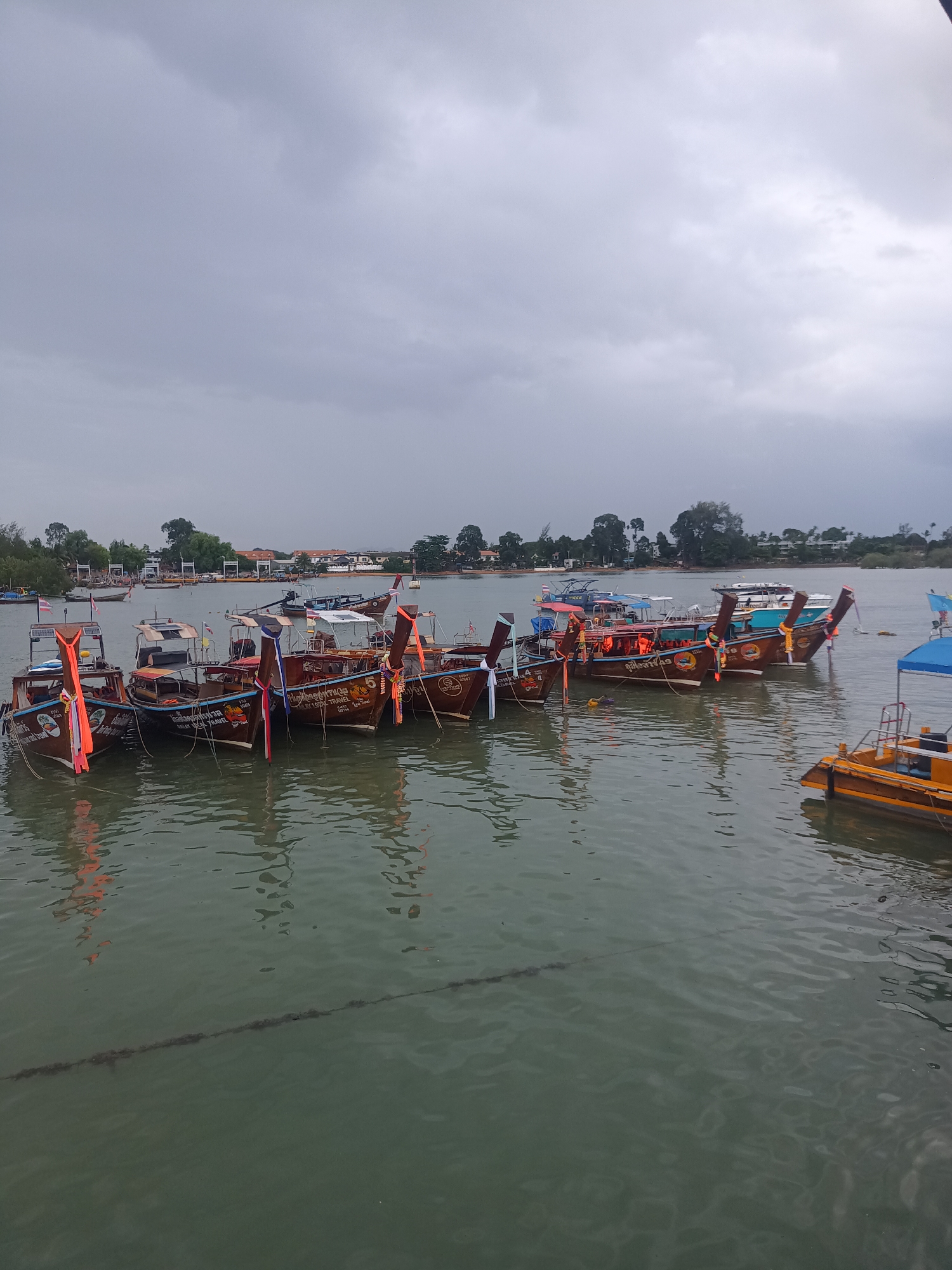 The image shows a harbor in Ao Nam Mao, Thailand, filled with parked longtail boats. The boats are primarily wooden, with decorative prows and colorful ribbons. Some boats are painted with numbers and text in Thai. A small, yellow and orange boat is visible on the right side of the frame. The water is a murky green color, and a mooring rope stretches across the foreground. The sky is overcast and gray. Buildings and trees are visible in the background across the water.