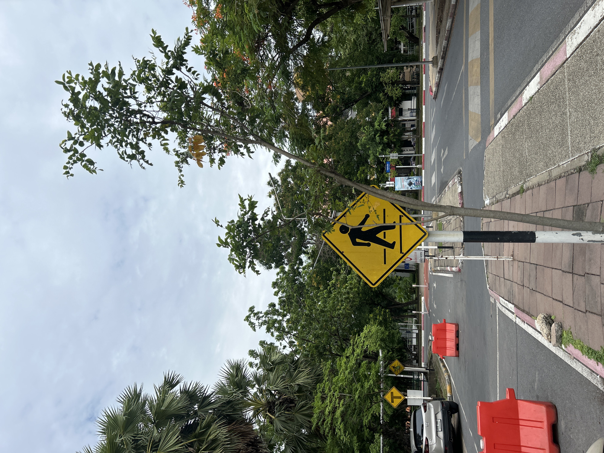 A diamond-shaped yellow pedestrian crossing sign with a black silhouette of a person walking is prominently displayed in the foreground. The sign is mounted on a black and white striped pole. To the left of the sign, a light-colored car is parked on the side of the road. Red plastic barriers are placed on the road, demarcating an area. A paved sidewalk with brick edging runs alongside the road. In the background, a street with marked lanes and a crosswalk is visible, lined with trees and some buildings. The sky is overcast. The scene appears to be in a town or municipal area, possibly a park or university campus, given the presence of pedestrian crossings, barriers, and greenery. Based on the provided information, this scene is located in Tha Khlong Town Municipality, Thailand.