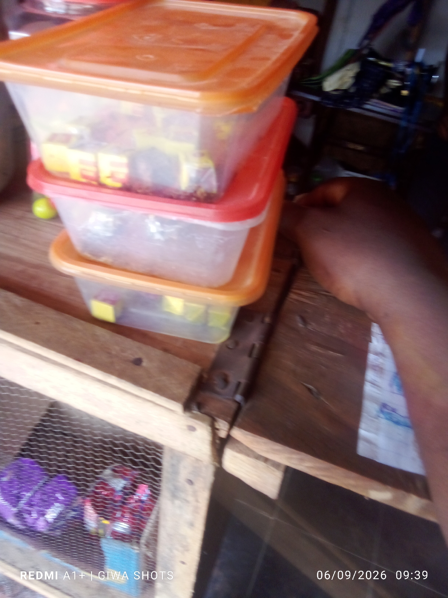 The image depicts a close-up of stacked plastic containers filled with various items, likely food products. The containers are translucent and some have colored lids (orange and red). Inside, yellow cubes, possibly bouillon cubes, are visible in the upper containers, and there are red and purple items in a wire mesh enclosure below. A person's arm and hand are visible on the right side of the frame, partially obscuring a piece of paper. The scene appears to be on a wooden surface. The timestamp "06/09/2026 09:39" and "REDMI A1+ | GIWA SHOTS" are present in the lower portion of the image, suggesting it was taken with a Redmi A1+ phone by "GIWA SHOTS" on September 6, 2026, at 09:39. The background is blurred but suggests an indoor setting with some shelving and miscellaneous items. Without further information or context, the specific location in Jalingo, Nigeria, cannot be determined from the visual elements alone.