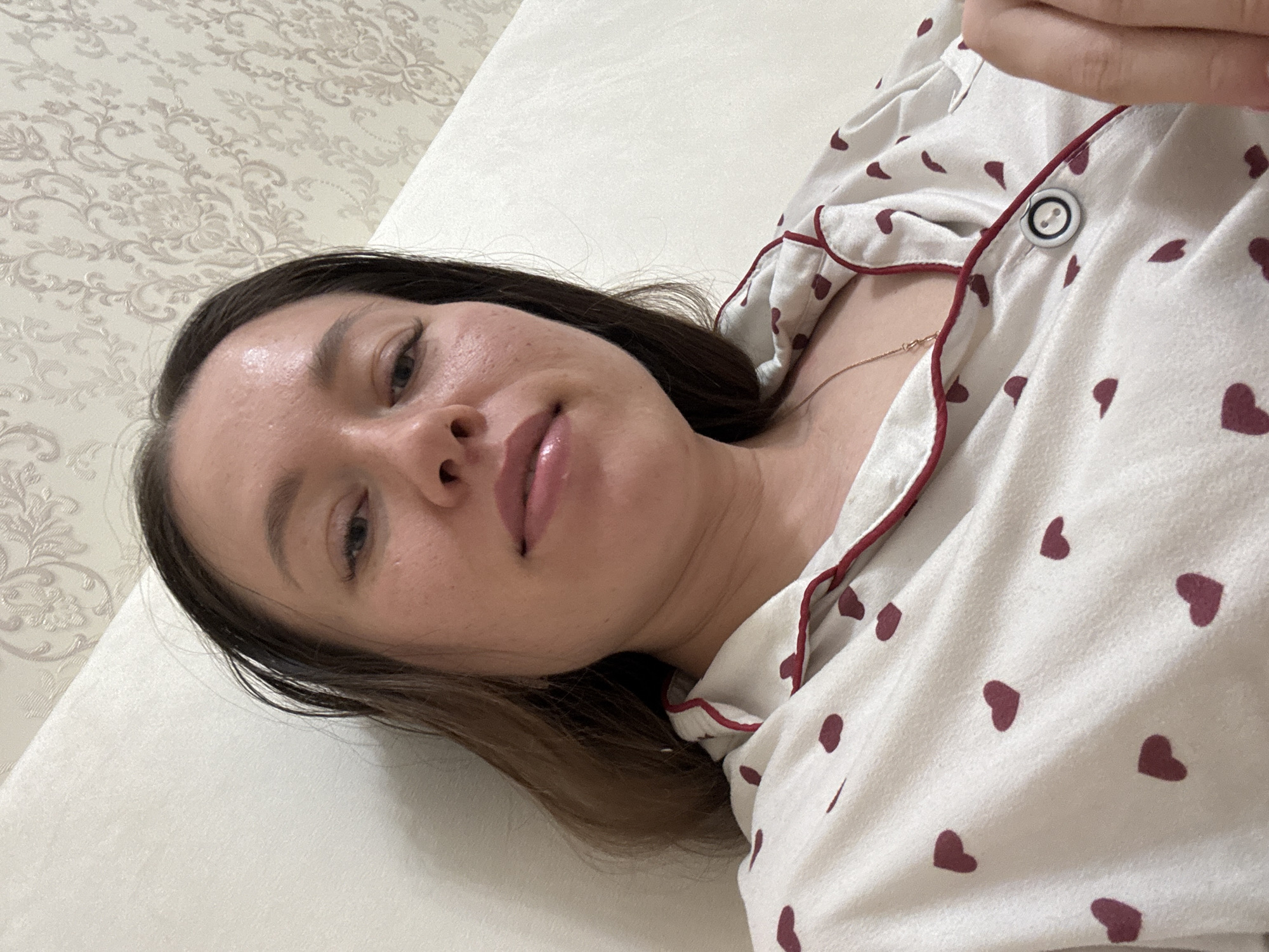 A woman is lying down, possibly on a bed, based on the plain white surface beneath her. She is wearing pajamas patterned with small, burgundy-colored hearts and has dark hair. The room is in Ферма, Russia, based on the image context. The wall behind her head features a patterned, floral wallpaper. A gold necklace is visible around her neck, above the pajamas. Her face has a dewy look.