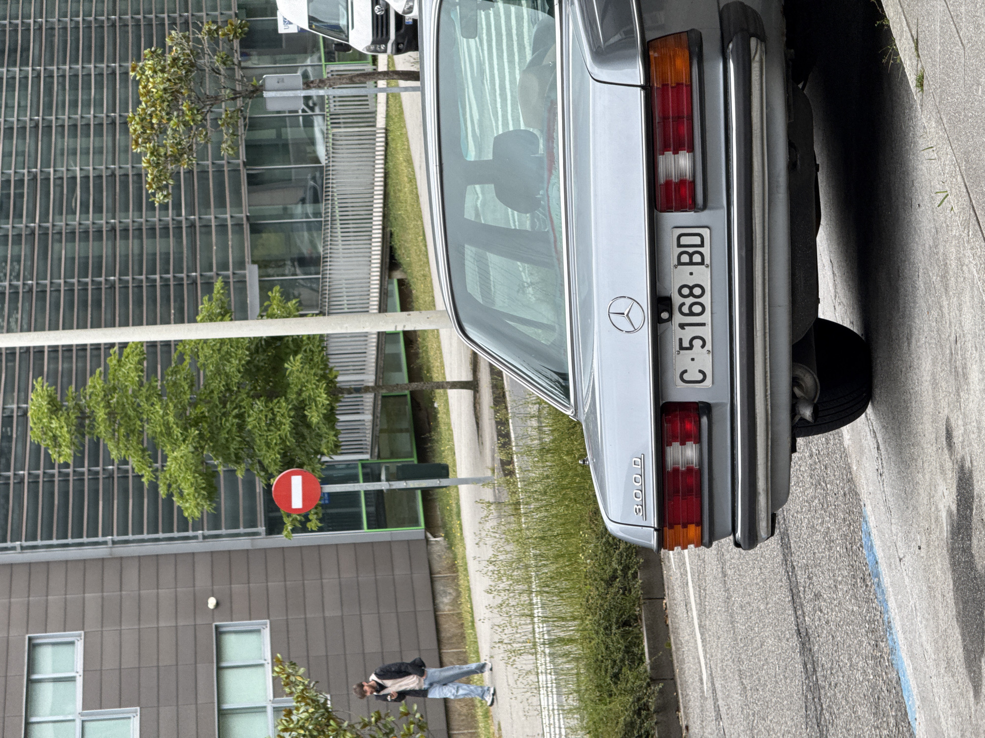 The scene depicts an outdoor urban environment in Santiago de Compostela, Spain, as indicated by the "C" prefix on the license plate. In the foreground, the rear of a light metallic blue Mercedes-Benz 300 D sedan is parked on the right side of an asphalt road. The vehicle displays a Spanish license plate reading "C-5168-BD". Its taillights feature amber, red, and clear segments, and a chrome bumper with black protective strips is visible. A single exhaust pipe extends from beneath the rear on the driver's side. The car's right rear tire is partially visible, and it is parked with a blue line marking painted on the curb next to it.
On the concrete sidewalk to the left of the car, a male pedestrian, estimated to be in his 20s or 30s with a beard, walks away from the camera. He is wearing a dark jacket over a light-colored top, light blue jeans, and white sneakers, and is looking down at a mobile phone held in his right hand. The sidewalk is bordered by a strip of green foliage, including grass and small bushes.
In the mid-ground, a red circular "No Entry" sign with a white horizontal bar is mounted on a grey pole. Behind this, a tall, white cylindrical pole stands prominently.
Two distinct buildings form the background:
1. On the left, a multi-story, dark grey building with a grid-like facade and rectangular windows with light frames.
2. On the right, a more modern, taller building featuring a glass facade with numerous horizontal louvers or sun-shading elements. The ground floor of this building includes green-framed window sections.
Further in the background, behind a metal fence, parts of two white vehicles are visible: a white panel van, likely a Volkswagen or Mercedes-Benz Sprinter, and a smaller white car. Several trees with green foliage are scattered throughout the scene, including a tall, slender tree near the "No Entry" sign and a broader tree with smaller leaves to its right. The lighting suggests an overcast or diffused sky.