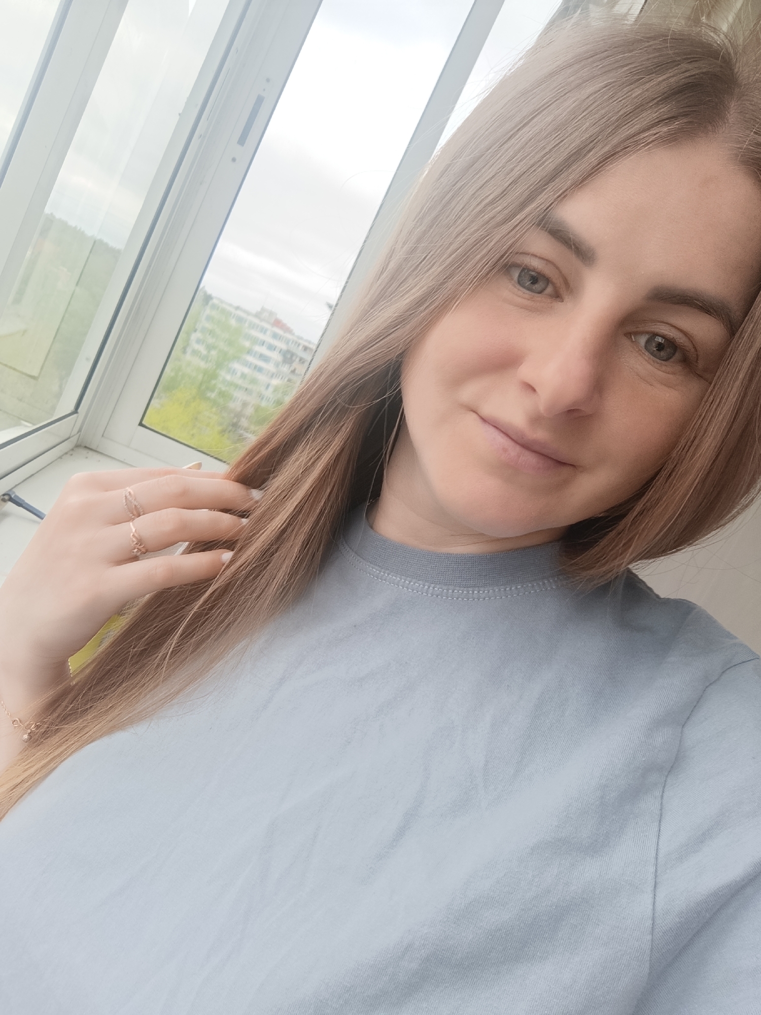 A woman with light brown hair is pictured in a selfie, wearing a light blue t-shirt. She is wearing two rings on her left hand. The background shows a window with a view of a city, including buildings and trees.