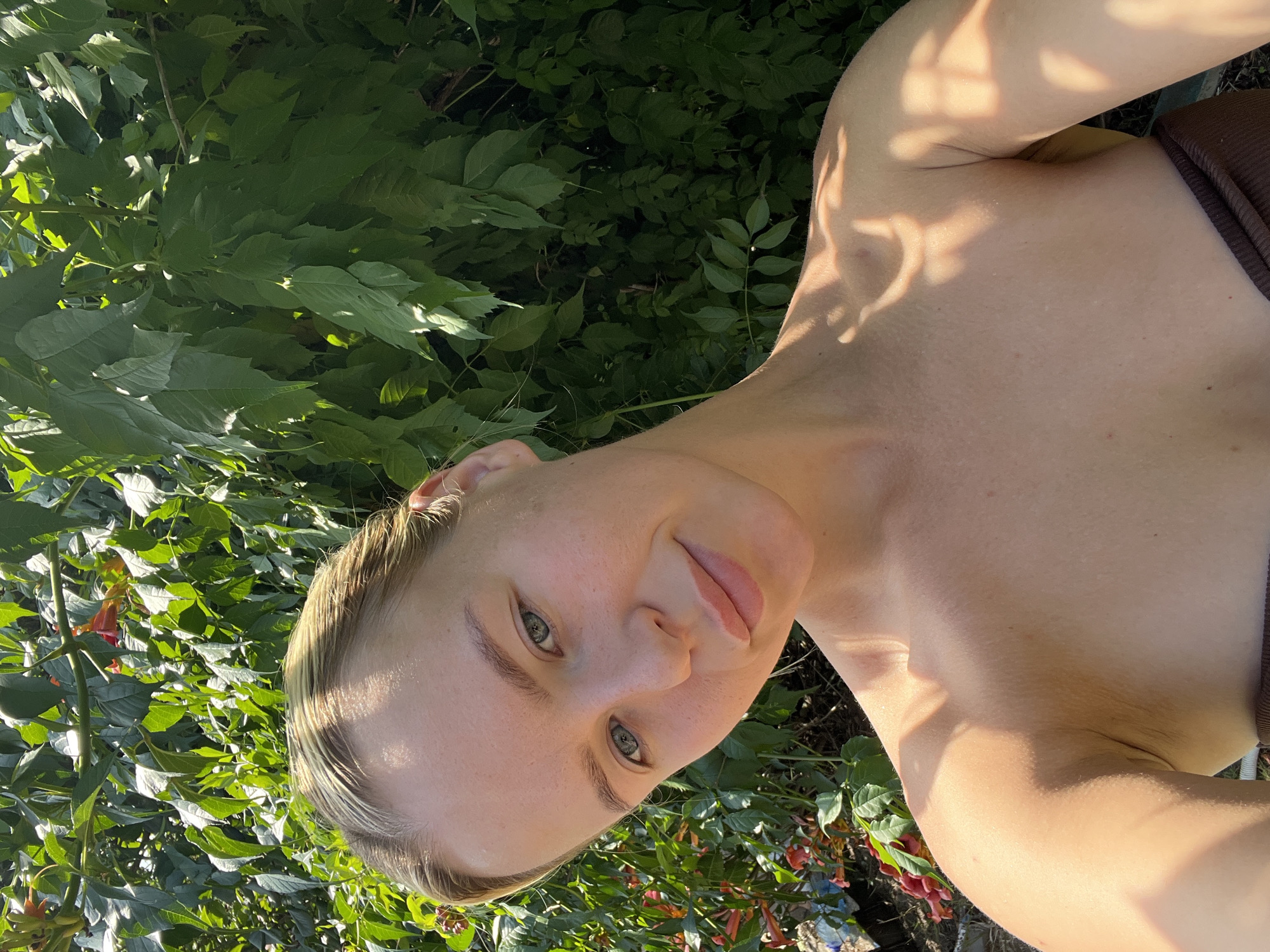 A young woman with blonde hair and blue eyes takes a selfie outdoors. She is looking at the camera with a slight smile. The background is a lush green bush with some red flowers. The sun is shining, casting shadows on her face and shoulders. She is wearing a brown top. The location is unknown.