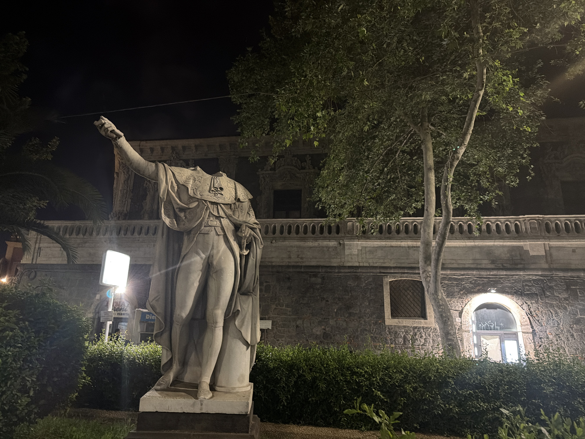 The image depicts a headless statue in the Piazza Vincenzo Bellini in Catania, Italy. The statue is of a man in classical garb with his right arm raised. It stands in front of a stone building with a row of arches along the top and two arched windows, one illuminated. Lush greenery surrounds the base of the statue and the building. A street lamp illuminates a sign to the left of the statue. The scene is at night.