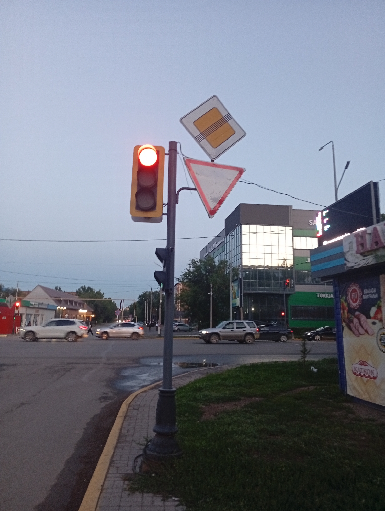 An outdoor, low-angle, full shot captures a street scene at dusk in Oral, Kazakhstan. A traffic light with a red illuminated signal stands prominently in the foreground, mounted on a grey pole. Above it, two road signs are affixed: a yellow diamond-shaped sign with black stripes and a red-bordered triangular sign pointing downwards. The street is paved, with a curved sidewalk made of brick and bordered by yellow paint. Cars, including a white SUV and a dark sedan, are visible on the road, with some appearing blurred due to motion. A pedestrian walks across the street in the distance. In the background, modern buildings with glass facades and illuminated signage, including "SA" and "TÜRKIA", are present. Greenery and trees line the street. A large advertisement banner featuring images of sausages and the text "KAZKON" is visible on the right side of the frame. The sky is a pale blue, indicating twilight.