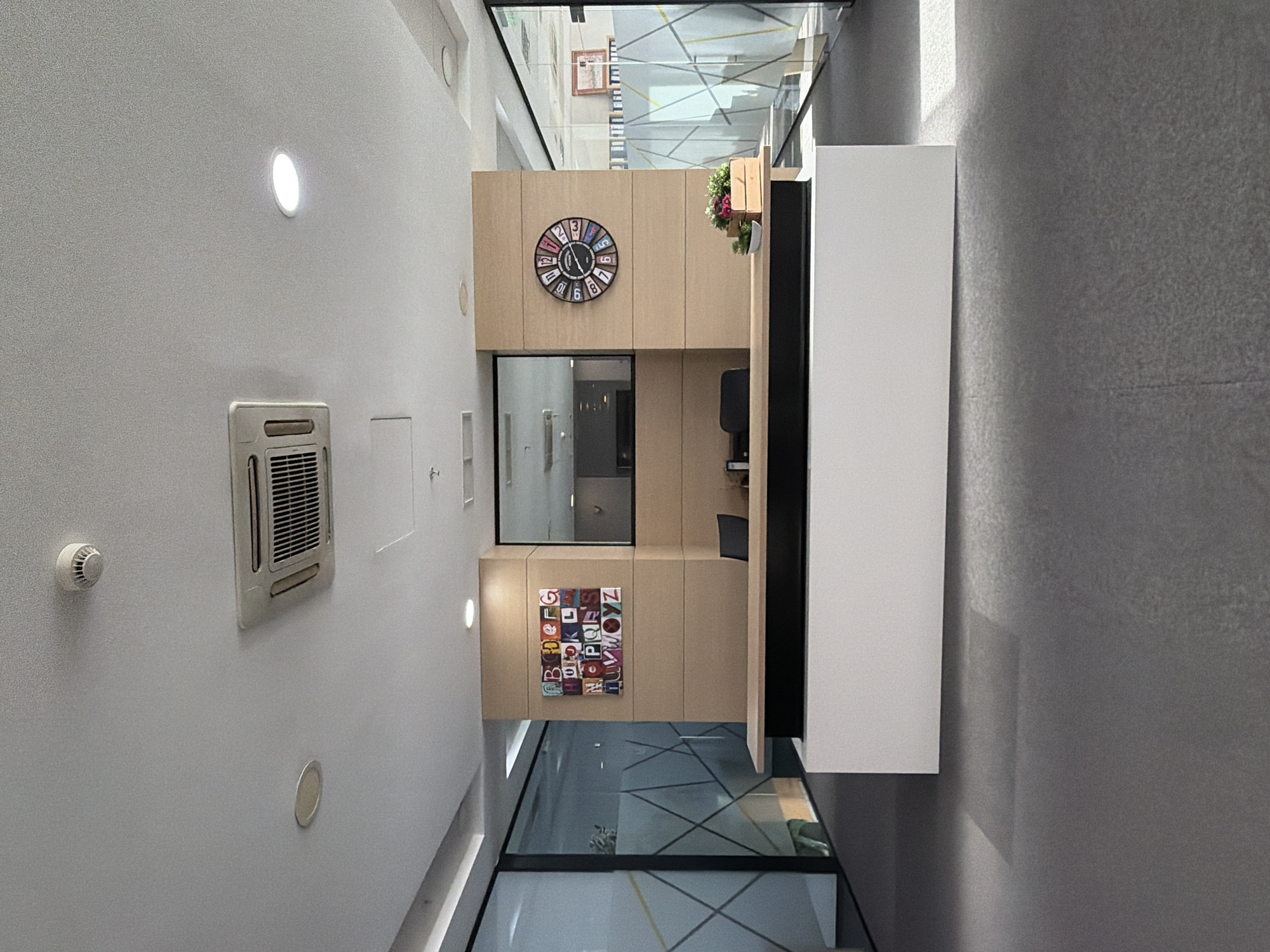 The image depicts an interior space with a modern design. A light-colored wall with an air conditioning unit, a smoke detector, and several small recessed lights is visible. Adjacent to this wall, there is a wooden structure containing a window, a clock, and an alphabet display. A white cabinet is positioned next to the wooden structure. Above, a glass ceiling allows a glimpse of the exterior, suggesting an open or atrium-like space. The flooring appears to be glass. The location is Sofia, Bulgaria.