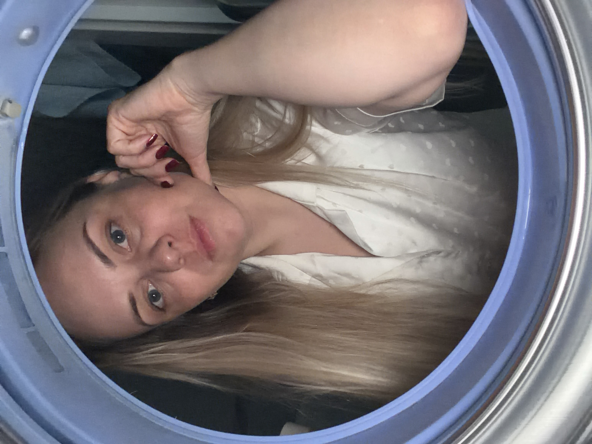 A fair-skinned woman with blonde hair and blue eyes is looking directly at the camera through the open door of a front-loading washing machine. She is wearing a white pajama top with a grey pattern. Her left hand is touching her face. There is clothing visible in the washing machine behind her. The scene is brightly lit. There is no indication of the location being Votkinsk, Russia.
