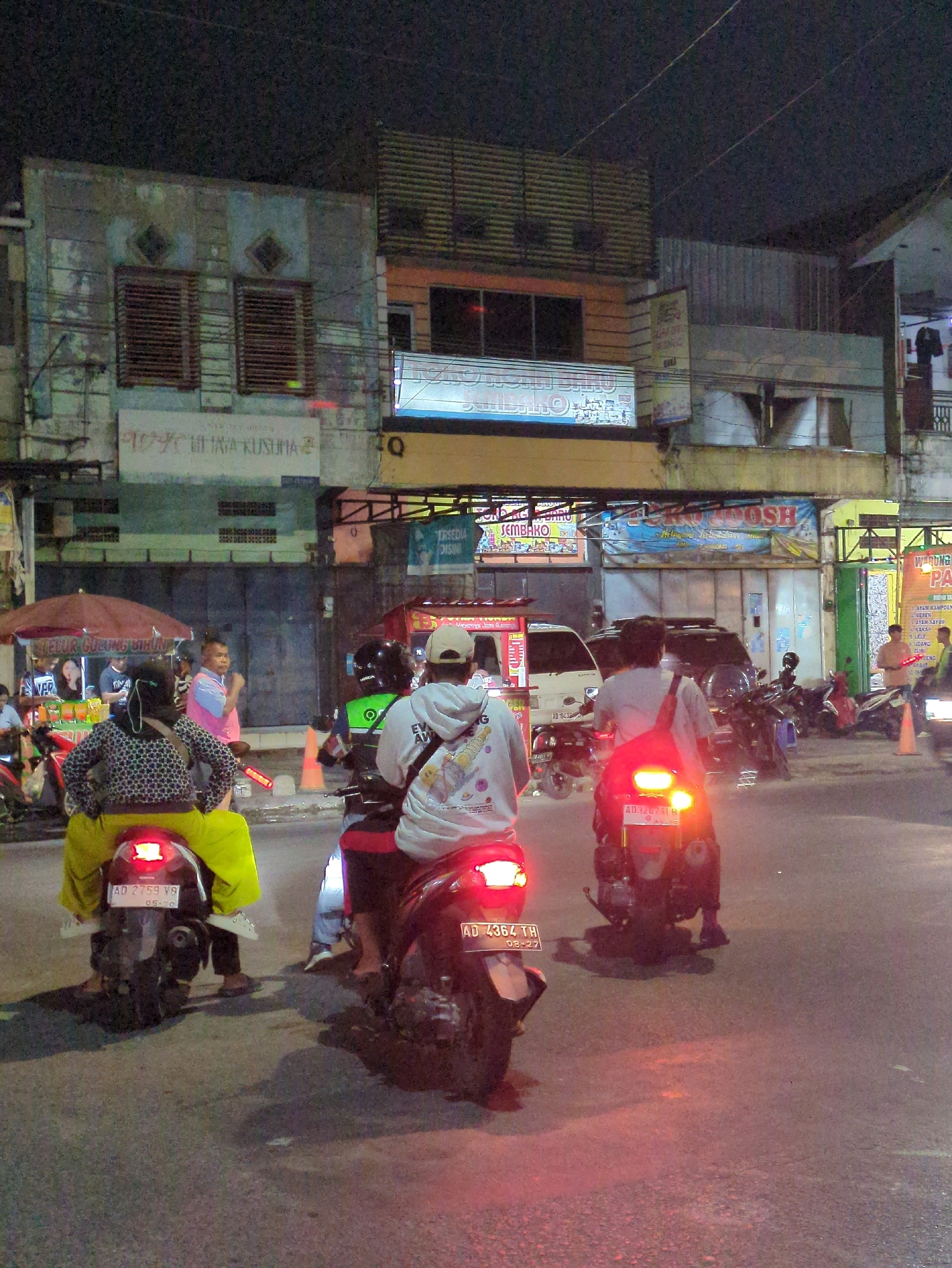At night on a city street in Gumpang, Indonesia, several people are riding motorcycles. In the foreground, two motorcycles are stopped in traffic, their taillights glowing red. The rider of the left motorcycle is wearing a black and white patterned top and yellow pants. The rider of the right motorcycle is wearing a white hoodie and jeans. In the background, there are buildings with illuminated signs, a food cart with a red umbrella, and several other motorcycles and vehicles. The street is paved and appears wet. The overall atmosphere is one of a busy urban environment at night.