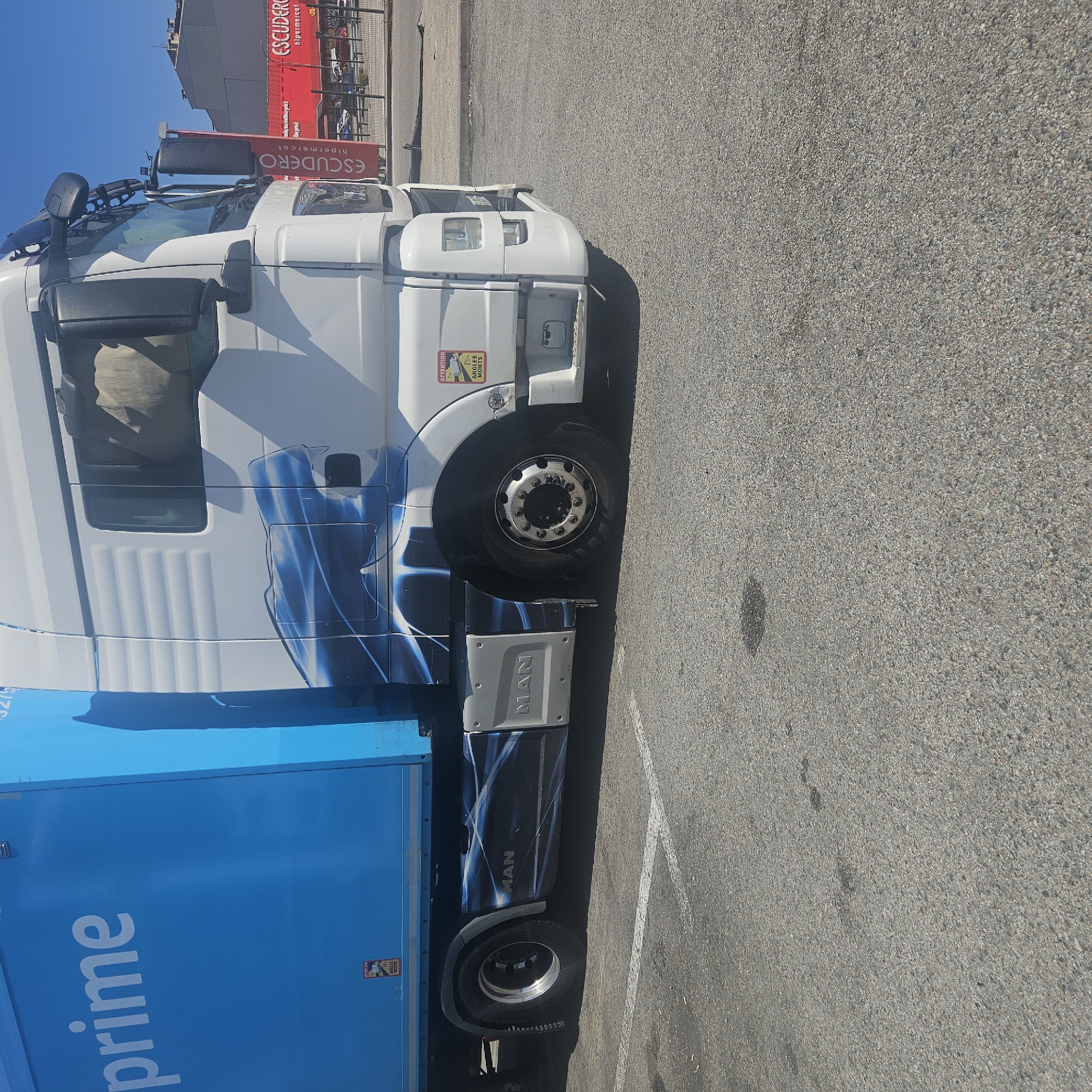 A white and blue Amazon Prime delivery truck is parked in a parking lot in La Jonquera, Spain. The truck has an MAN logo on the side and "prime" printed in large white letters on the blue cargo box. The truck is parked on a grey asphalt surface, which is marked with a white parking line. In the background, a red building with the sign "ESCUDERO hipermercat" is visible, indicating the location in Spain. The sky is blue and clear. There are "Attention Angles Morts" stickers on both the cab and cargo box of the truck.
