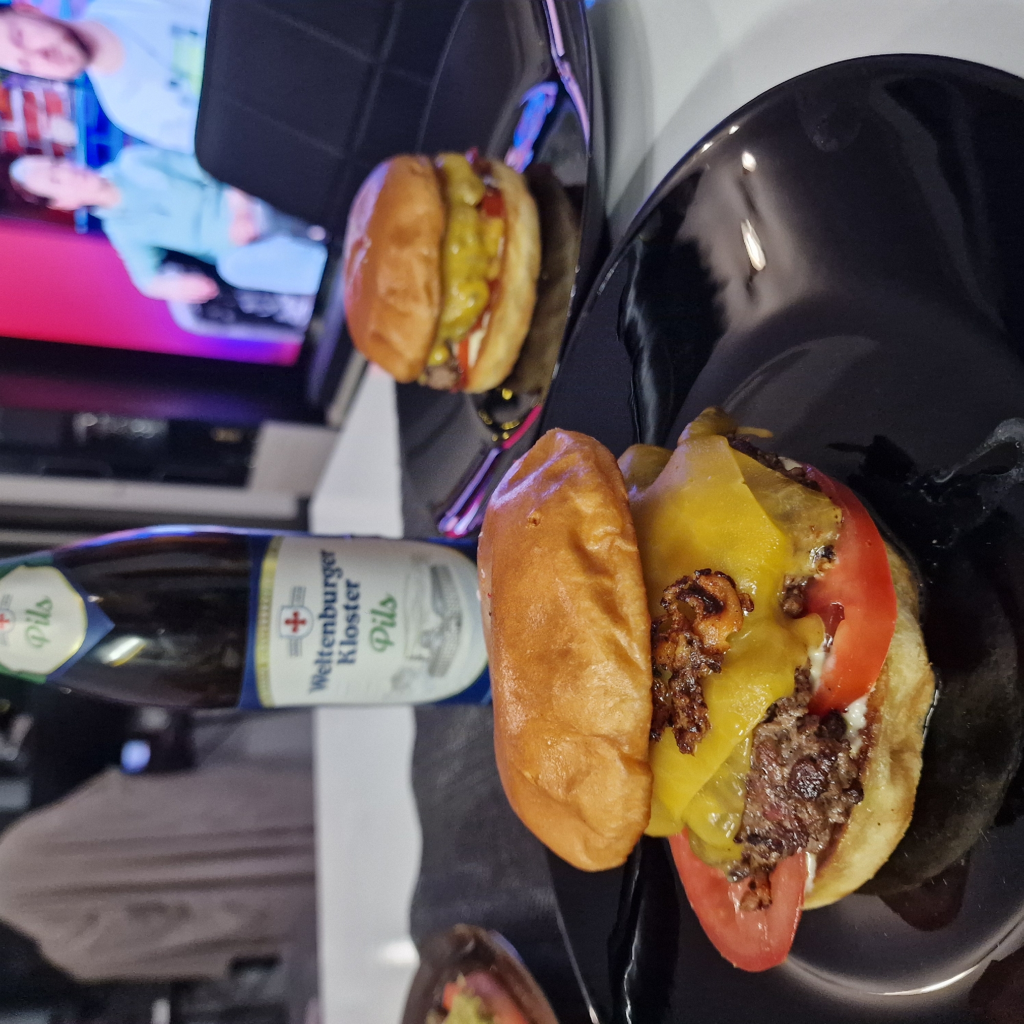 The image shows two hamburgers on black plates, next to a bottle of "Weltenburger Kloster Pils" beer. The hamburgers appear to be homemade and contain beef patties, cheese, tomatoes, and onions. They are served on toasted buns. The beer bottle is dark green and has a white label. The background shows a television screen and a white surface. There is no evidence in the picture to indicate that this photo was taken in Sofia, Bulgaria.