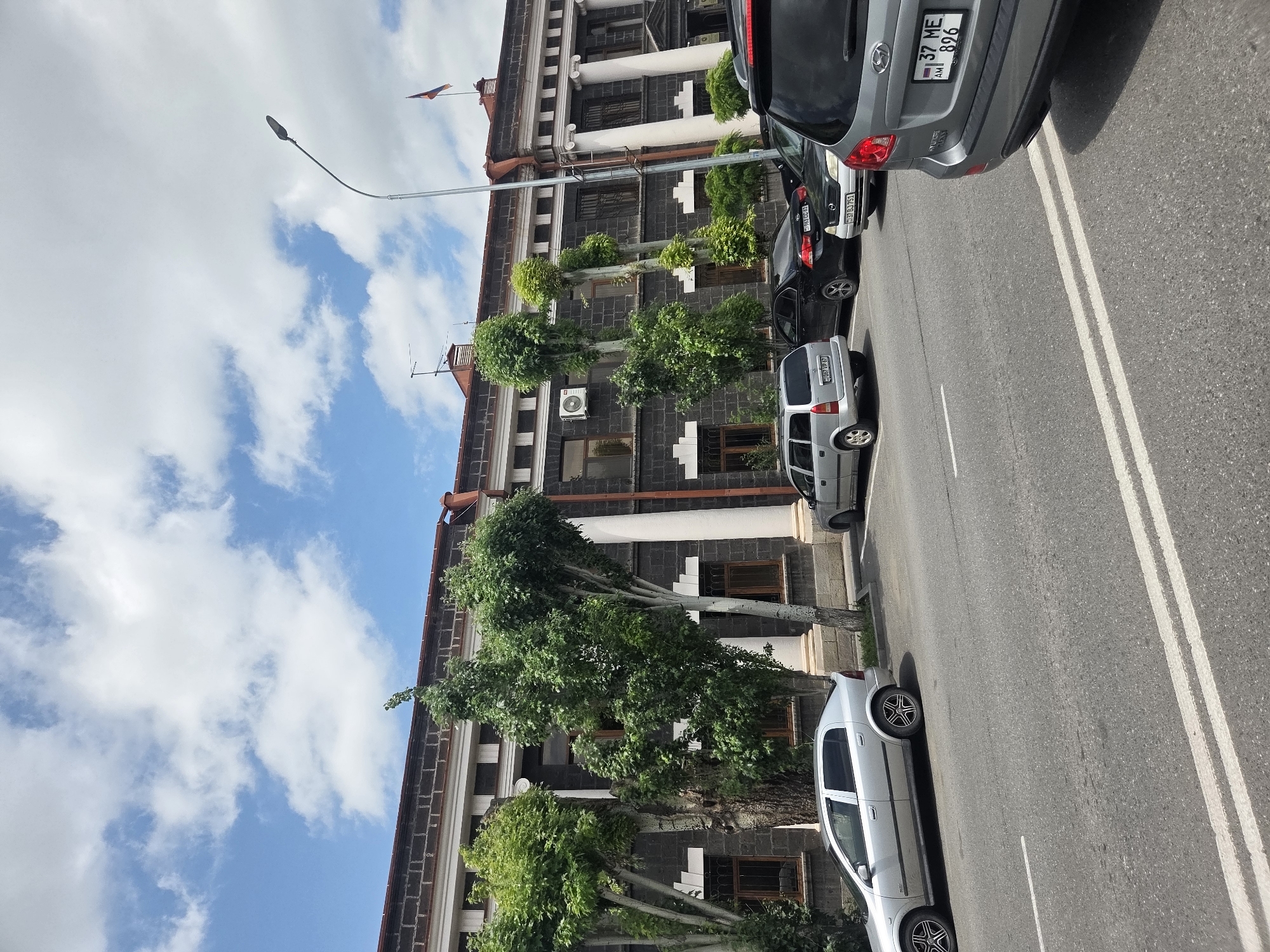 The image shows a street scene in front of a large, dark-colored building with white columns. The building appears to be in an urban environment, with cars parked along the street and in front of the building. Trees with green foliage are planted in front of the building. The sky is blue with scattered white clouds. The building has a red roof and several windows. A street light is visible on the right side of the image, and an Armenian flag is flying from the top of the building. The license plates of some of the cars are visible, indicating they are from Armenia. One car, a silver sedan, is parked on the left side of the street, facing left. Another silver SUV is parked to its right, facing right. A black sedan is parked behind the SUV, and a gray SUV is parked on the far right, facing right. The road in the foreground has double white lines. The overall scene suggests a typical day in a city in Armenia.