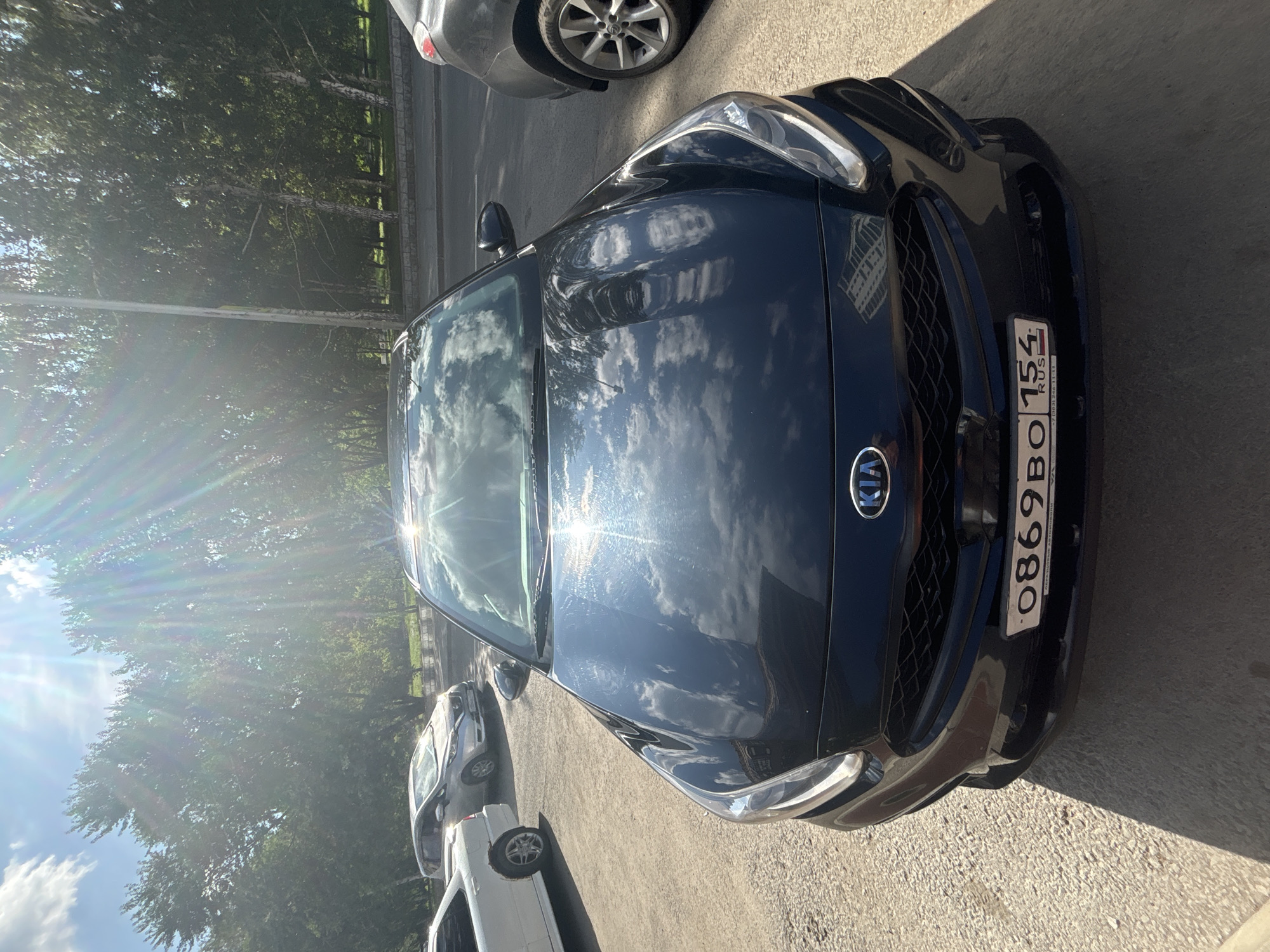 A black Kia sedan is parked on a paved surface in front of a line of trees. The car's front end is visible, including its grille, headlights, and license plate. The license plate reads "0869BO 154 RUS". The sun is shining brightly, casting shadows and creating lens flare in the image. In the background, other cars are parked and trees line a street. The environment appears to be an urban setting.
