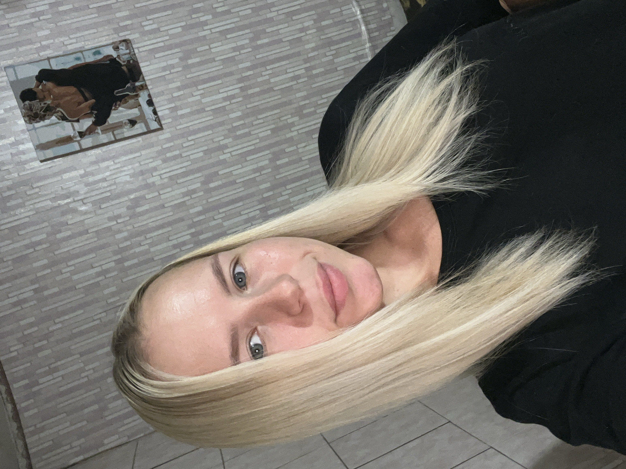 A close-up, medium shot shows a young woman with blonde hair posing for a selfie in a room. She has fair skin and blue eyes, and she is wearing a black top. Her hair is straight and falls past her shoulders. The background consists of a wall with a patterned wallpaper. On the wall, a small framed picture of a couple embracing is visible. The lighting is soft, illuminating the woman's face and hair. The overall scene appears to be indoors.