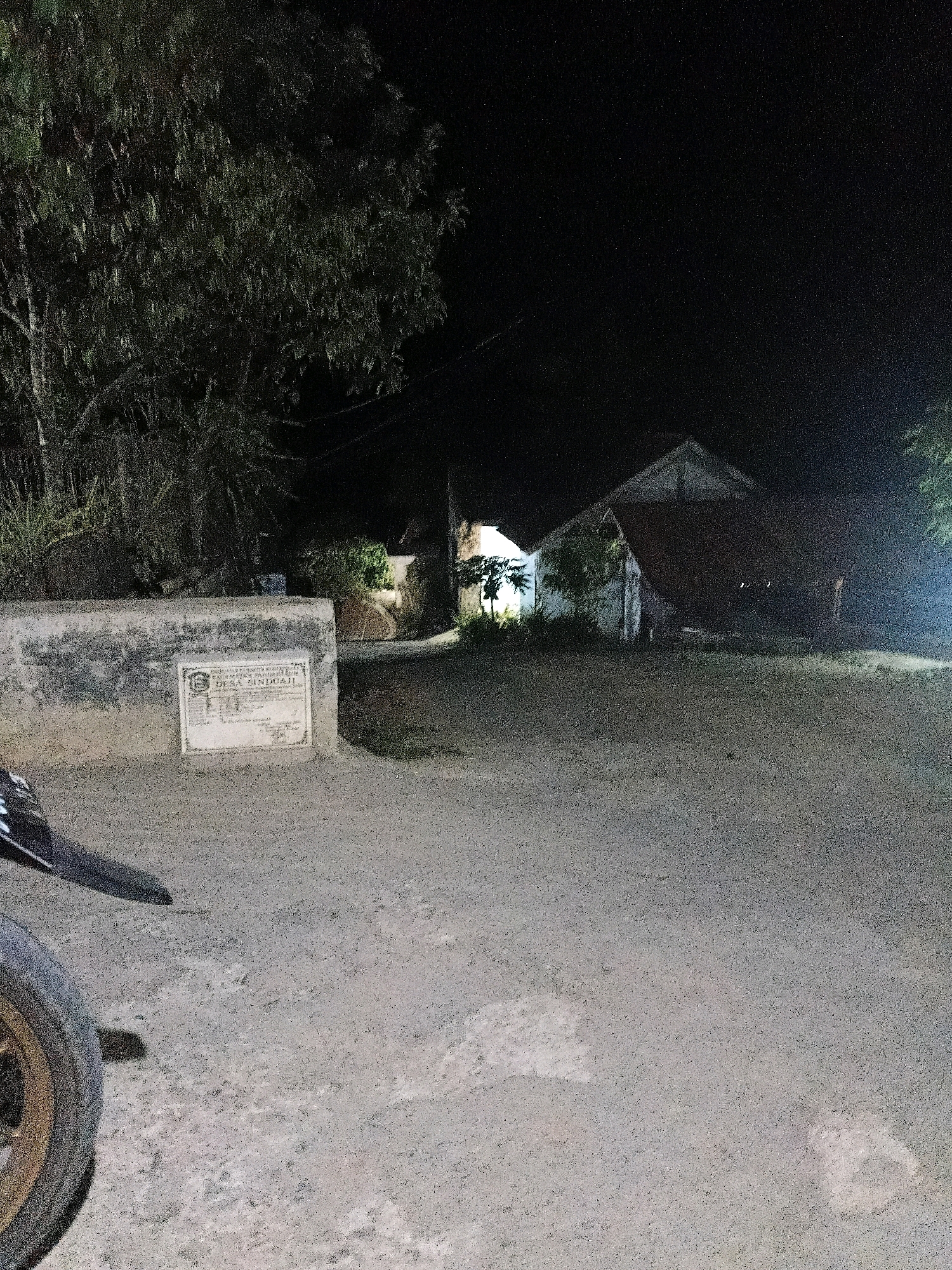 The image depicts a nighttime scene in the city of Sinduaji, Indonesia, characterized by a rural or suburban environment. In the foreground, a dirt or gravel road stretches towards the background, illuminated by artificial light, likely from a nearby source not visible in the frame. To the left, the front portion of a motorcycle is visible, including a tire with a metallic rim and a section of the frame. Adjacent to the road and slightly elevated is a low, concrete wall, likely part of a building or boundary. Attached to this wall is a plaque with text and a crest, which appears to be an official marker or sign for the area, possibly indicating the name of the village or a historical monument.
Behind the wall, trees and foliage are visible, their shapes obscured by the darkness. Further in the background, two simple, single-story houses are discernible. One house is partially illuminated, revealing white walls and a pitched roof. The other house is darker, with a prominent roofline. The overall impression is one of a quiet, residential area at night, with the presence of a motorcycle suggesting recent or ongoing activity. The lighting is dim, creating a slightly grainy texture in the image, but the key elements of the scene remain identifiable.