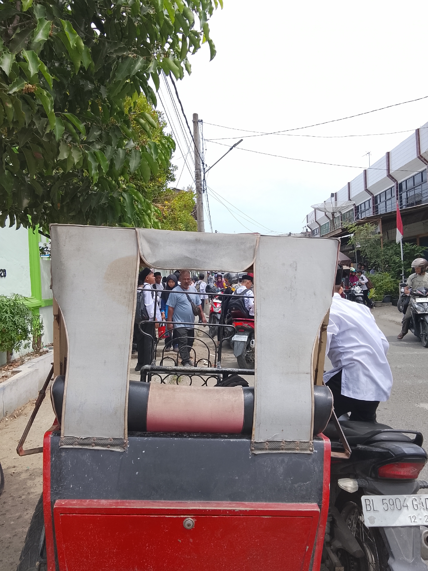 The image depicts a street scene in Lhokseumawe, Indonesia, likely during the day. In the foreground, a red and black carriage-like structure, possibly part of a pedicab or similar vehicle, dominates the lower portion of the frame. A person wearing a white shirt is seated on a motorcycle behind this structure, facing away from the camera. The license plate of the motorcycle reads "BL 5904 GAD".
In the midground, a group of people are visible. Some appear to be wearing school uniforms with white shirts and black pants, and some are wearing traditional Islamic caps. They are standing on or near the road, with several motorcycles parked around them. A man in a blue shirt is walking among them.
The background shows a street lined with buildings, some of which appear to be commercial establishments with multi-story facades. A red and white Indonesian flag is visible on one of the buildings. Overhead power lines and a street light are also present. Lush green trees with dense foliage line the left side of the street. The sky is overcast. The overall impression is of a busy urban environment with ongoing pedestrian and vehicular activity.