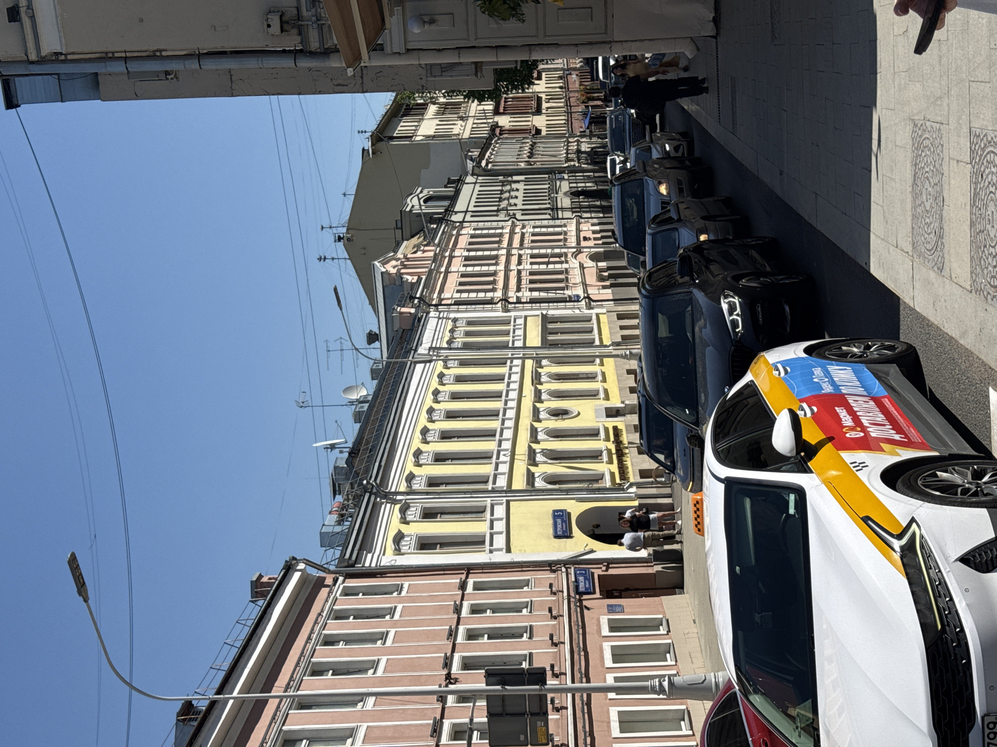 The image depicts a street scene in Moscow, Russia. The view is of a row of buildings on a sunny day. The buildings are multi-story with ornate facades, painted in yellow, pink, and off-white. A street sign for Petrovsky Street is visible. Cars are parked along the street, including a white taxi cab with a distinctive yellow and blue roof. A few pedestrians are visible on the sidewalk. Overhead, there are power lines.