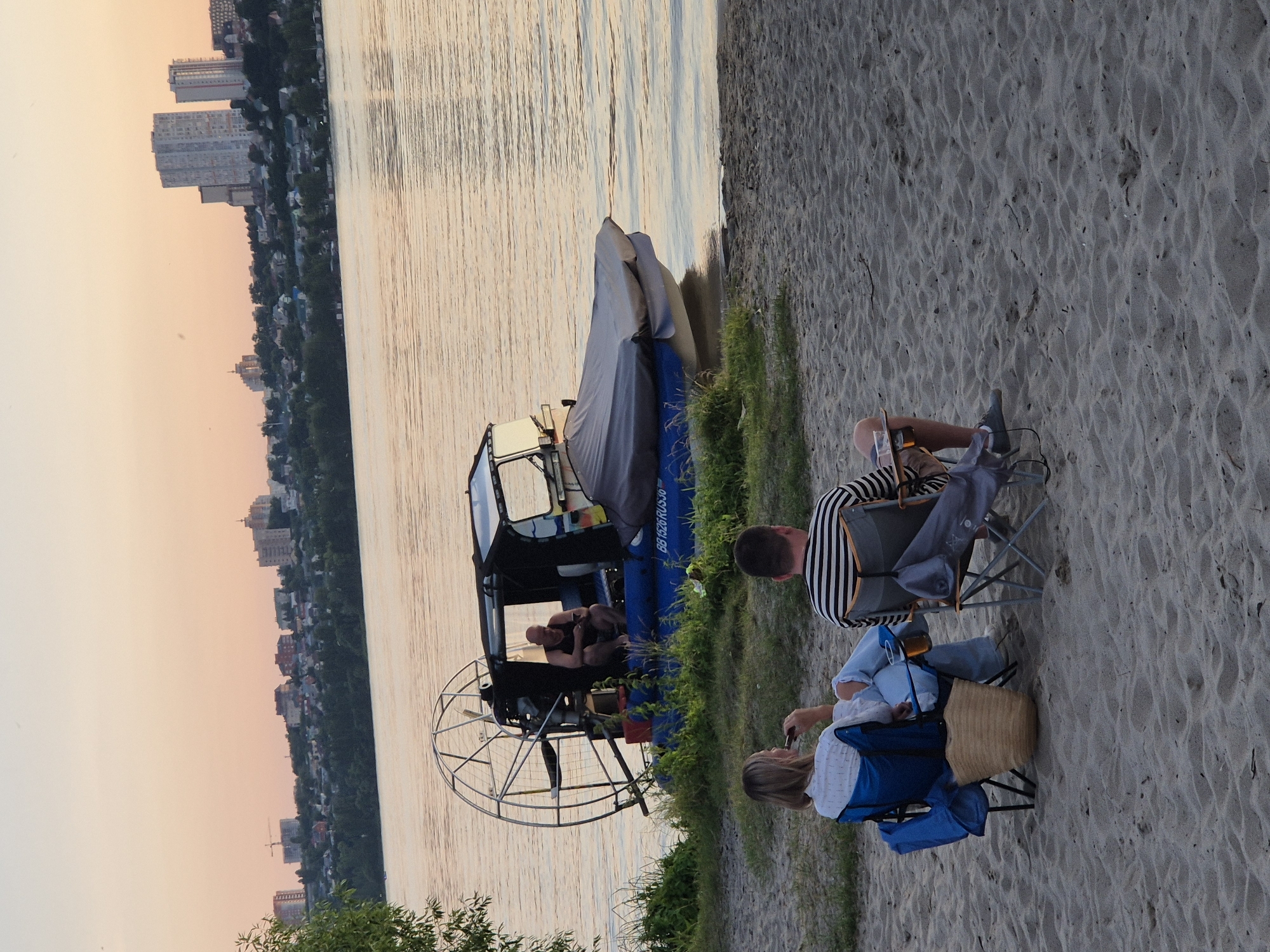 This is a vertically oriented, eye-level, wide shot taken outdoors on a sandy beach next to a body of water. The scene depicts a relaxed gathering with several people and an airboat. In the foreground, two people are seated in folding chairs on the sand. One individual, wearing a black and white striped shirt and shorts, is seated and looking away from the camera. Beside them, another person in a white shirt and blue jacket is seated and holding a cigarette. A straw woven bag is placed on the sand next to them.
In the mid-ground, an airboat is partially visible, pulled up onto the beach. It is covered with a gray tarp, and a blue panel is visible on its side. Inside the airboat's cabin, two people can be seen. The airboat has a large fan at the rear.
The background shows a body of water with gentle ripples, reflecting the soft light of the sky. Across the water, a city skyline is visible, featuring a mix of residential buildings and trees. The sky has a warm, hazy hue, suggesting either sunrise or sunset. The sand is textured with footprints. The overall atmosphere is calm and leisurely.
The visible text on the airboat reads "BB1526RUS50". The location is identified as Voronezh, Russia.