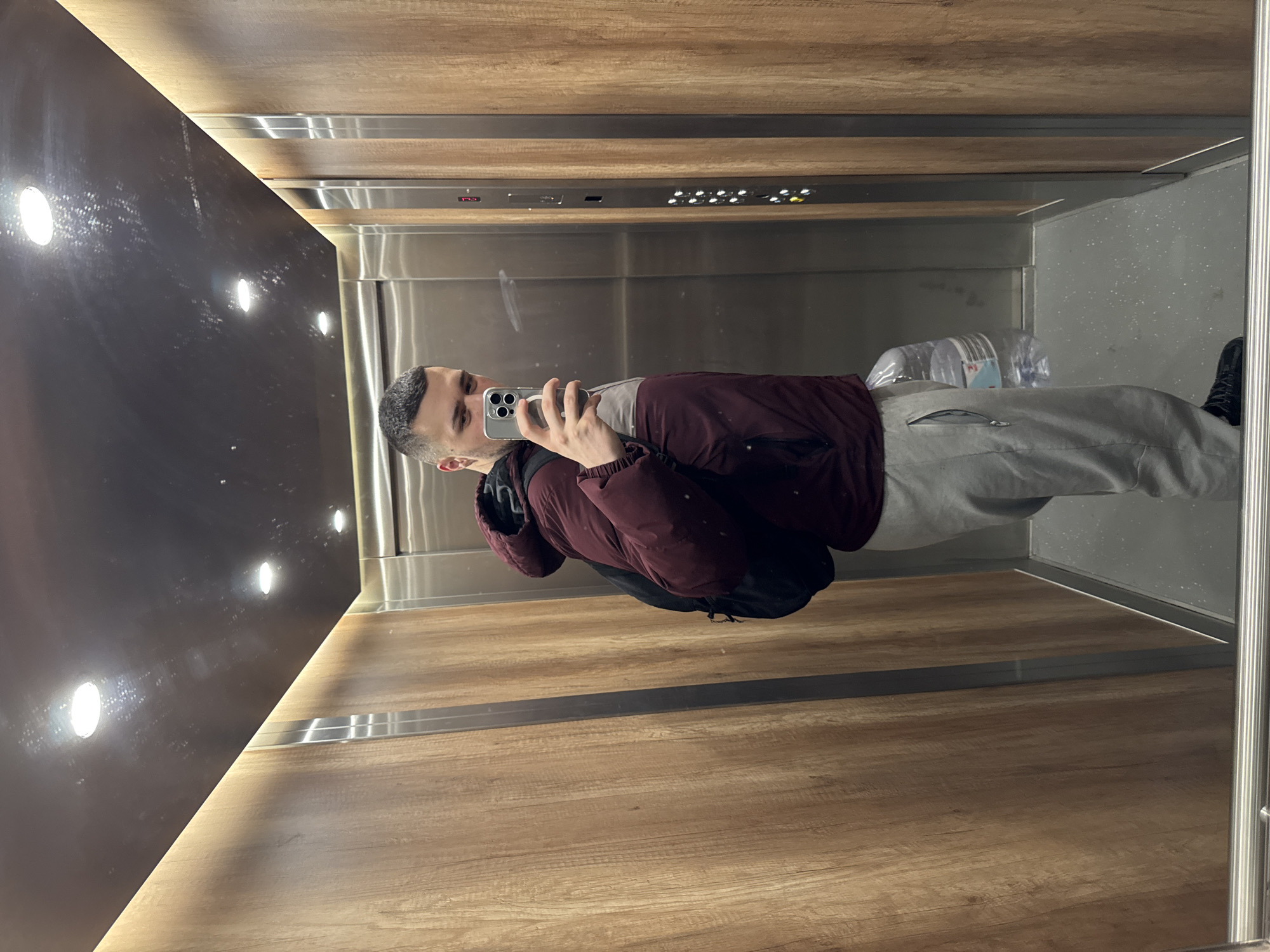 A man is taking a selfie in an elevator. He is wearing a maroon jacket, gray sweatpants, and black shoes. He is holding a smartphone in his right hand. Behind him, there are bottles of water on the elevator floor. The elevator walls are wood-paneled with a strip of buttons and lights. The scene appears to be inside an elevator. The location cannot be identified as Sofia, Bulgaria, based on the provided image alone.