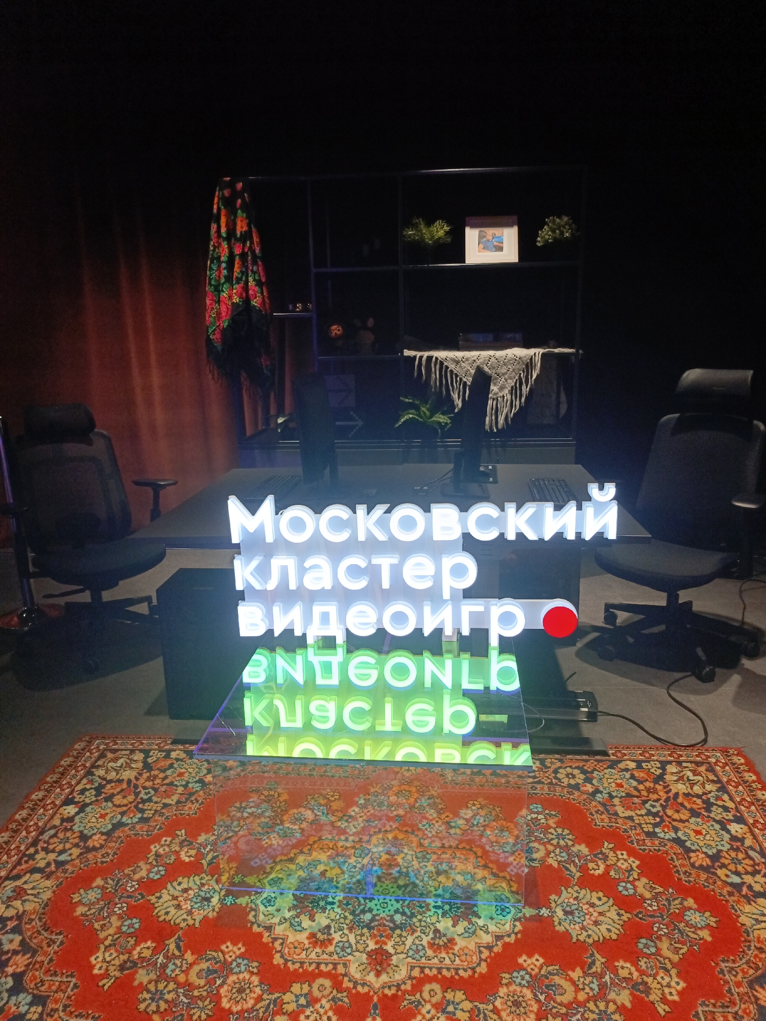 The image shows a room, likely indoors, in what is possibly a workspace or studio setting. Two black office chairs flank a long desk, each in front of what appears to be a computer monitor. Centered in front of the desk is a neon sign in Russian, which translates to "Moscow video game cluster." The sign's lighting casts a green and white glow on the immediate surroundings.
Behind the desk, there's a black shelf with various objects, including plants, a framed picture, and a crocheted throw. A red and floral patterned scarf hangs to the left of the shelf. A traditional Russian rug with an ornate pattern covers the floor in the foreground. The setting appears to be in Moscow, Russia, judging by the text on the neon sign.