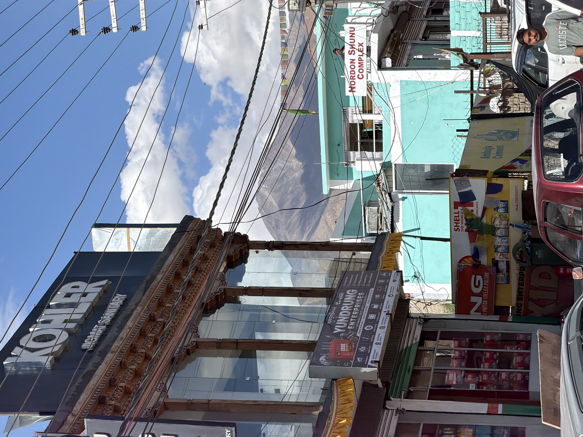 The image shows a street scene in Leh, India, with buildings lining a narrow road. On the left, a building with a dark facade displays a large "KOHLER" sign and a smaller "JIGMAT SANITARY" sign. Below this, the facade features ornate wooden carvings and large glass windows. A banner for "YUNDRUNG ENTERPRISES" is visible, advertising automobile spare parts and services. To the right, a bright teal-colored building has a sign for "NORDON SHUNU COMPLEX". A variety of electrical wires crisscross the sky, with prayer flags strung across them in the distance. In the foreground, several vehicles are parked, including a red car and a white car. A young man wearing a grey t-shirt with a "LACOSTE" logo stands near the white car. The background consists of rugged mountains under a blue sky with scattered white clouds. The overall impression is a bustling street in a mountainous region, likely during daytime.