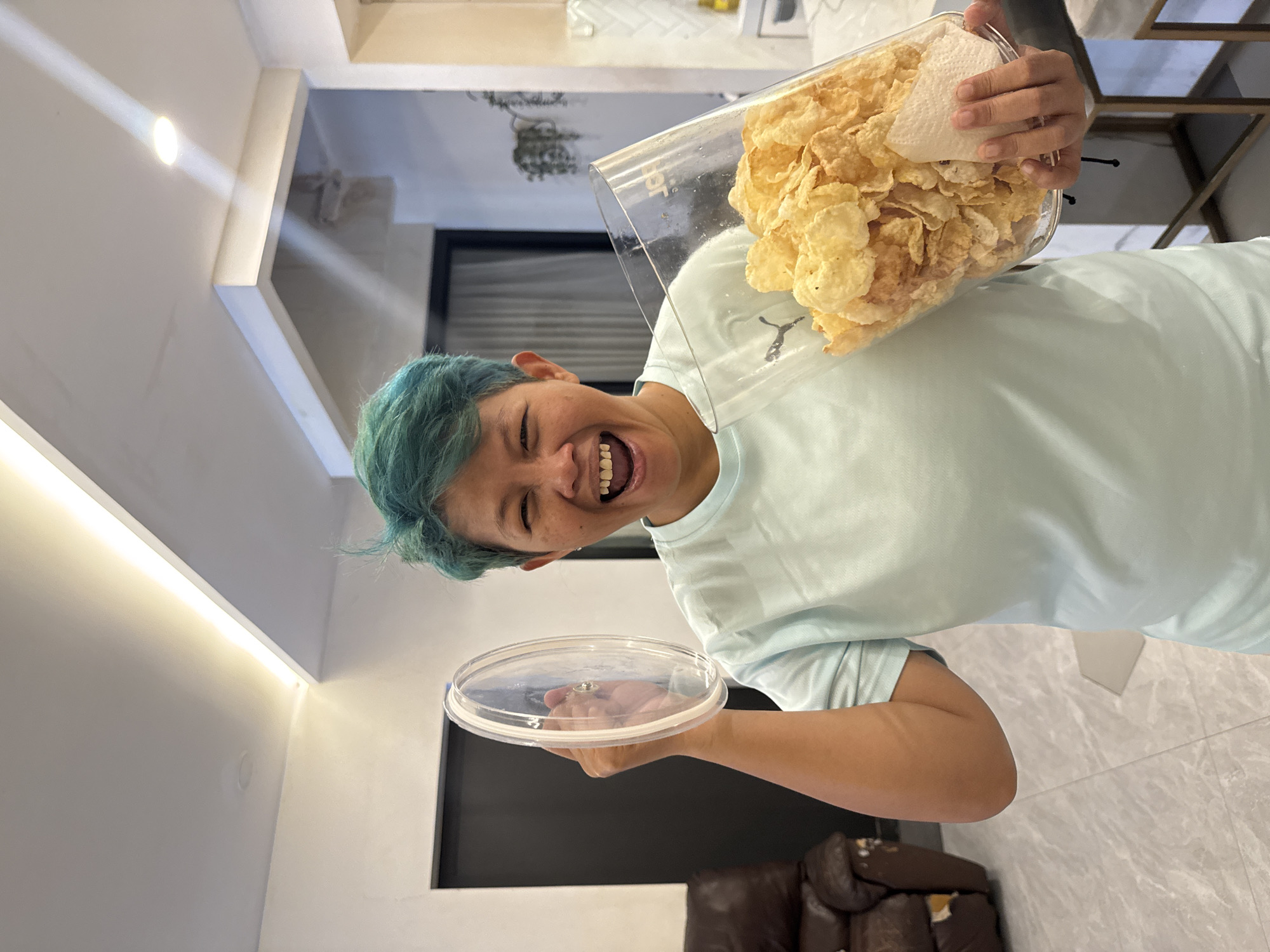 A person with short, turquoise-colored hair is holding a large, clear plastic container filled with corn flakes or similar chips. The container is open, and the person is smiling broadly, displaying their teeth. They are wearing a light blue T-shirt. In their other hand, they are holding the lid of the container. The background shows a modern interior setting, with white walls and ceiling, an LED strip light, a black door or panel, and a glimpse of a brown leather sofa. The location appears to be in a residential setting, possibly in the city of Bekasi, Indonesia, based on the interior design and general aesthetic. The image is oriented sideways.