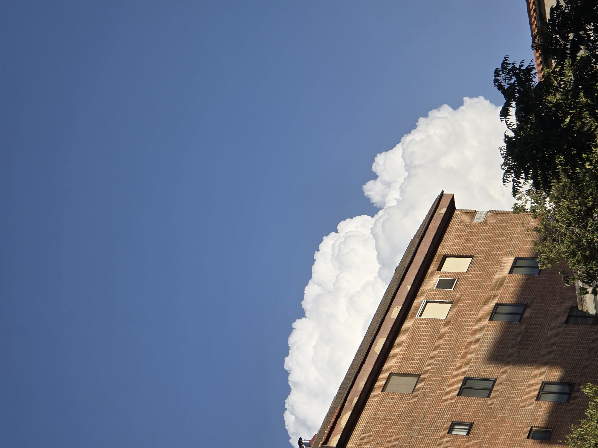 A brick building with numerous windows is seen against a bright blue sky, with a large, white cloud formation behind it. The building's roof is visible at the top left, and trees are in the foreground on the right. A shadow from an unseen object is cast across the building's facade.