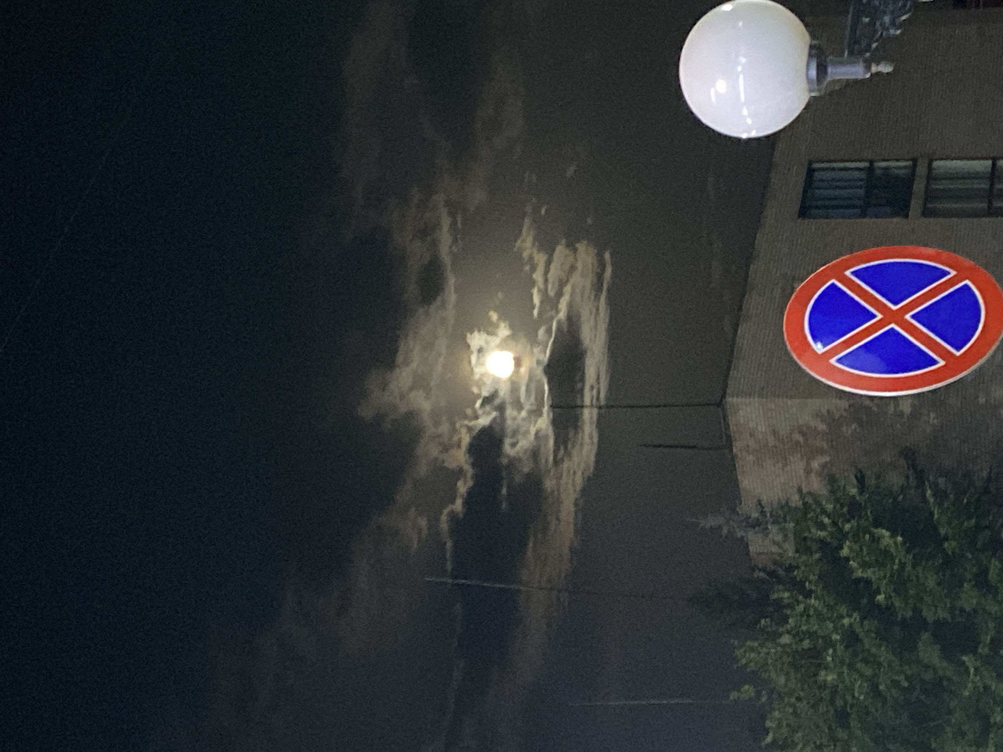 The image depicts a nighttime scene with the moon visible through clouds. In the foreground, there is a red and blue circular traffic sign with a blue "X" in the center, indicating "no stopping." To the right of the sign is a white spherical street lamp. Behind the sign and lamp, a brick building is visible, with trees to the left of the building. The overall scene suggests an urban or suburban environment. The image was captured in Ishimbay, Russia.