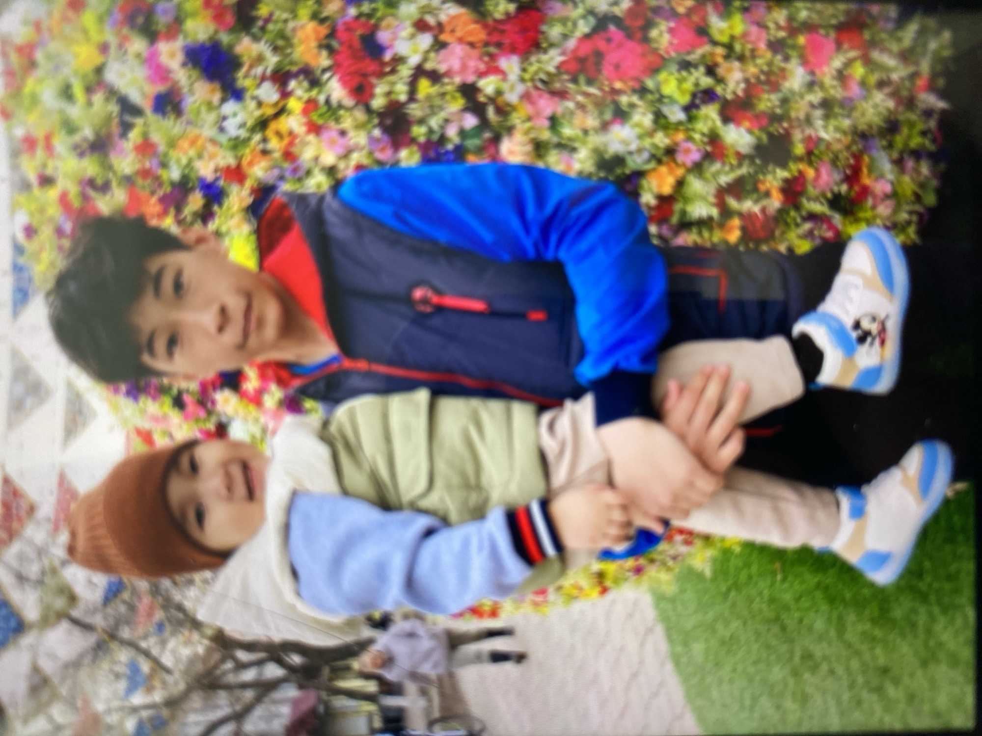 The image shows two young individuals, likely brothers, with the elder holding the younger in his arms. The elder boy, with dark hair, is wearing a blue and dark gray vest over a blue long-sleeved shirt with a red collar. The younger child, a toddler, is wearing a brown knit hat, a light-colored vest over a light blue garment, and beige pants. The toddler's shoes are white and blue with a Mickey Mouse design. They are standing in front of a vibrant, colorful floral arrangement. In the background, on the left side of the image, a person in a light-colored hoodie and pants is walking away from the camera. The ground in the foreground appears to be a combination of green artificial turf and patterned paving stones. The overall setting suggests an outdoor public space, possibly a park or a festival, within the Бескөл ауылдық округі, Kazakhstan.