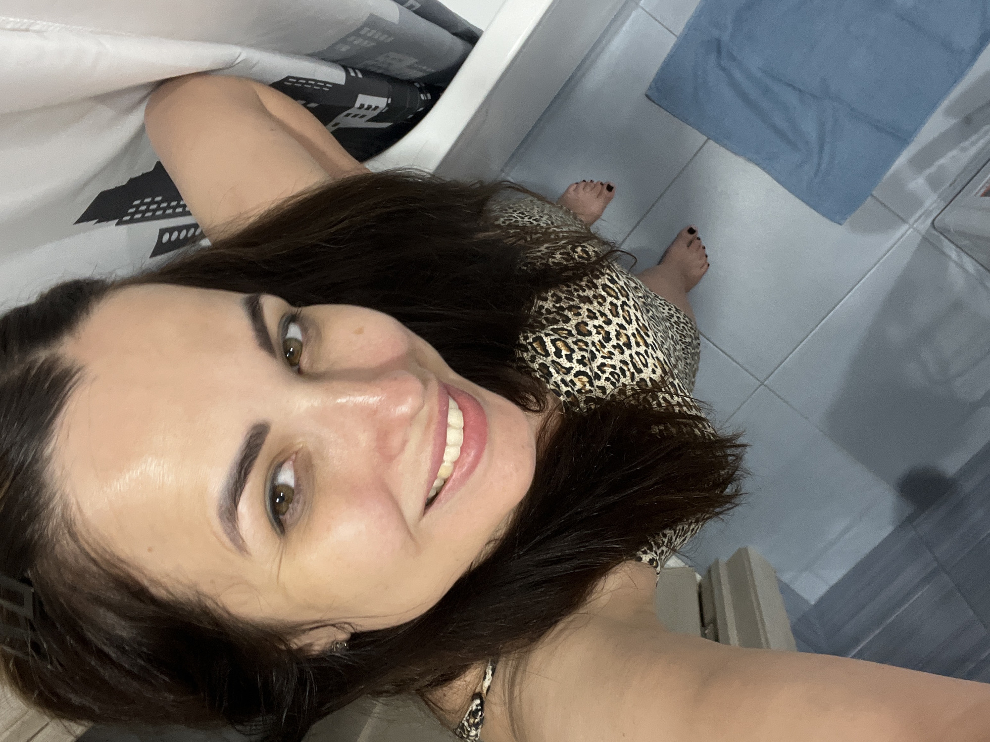 A woman with long, dark hair and brown eyes is captured in a selfie from a high angle in a bathroom. She is wearing a leopard-print dress and is positioned near a bathtub. The bathroom features light blue tiled flooring and a white bathtub. A grey and white shower curtain with a city skyline pattern hangs beside the tub. Her right arm is raised, and her left arm is bent. She is smiling at the camera. Her feet, with black nail polish, are visible on the tiled floor, and a blue towel is placed nearby.