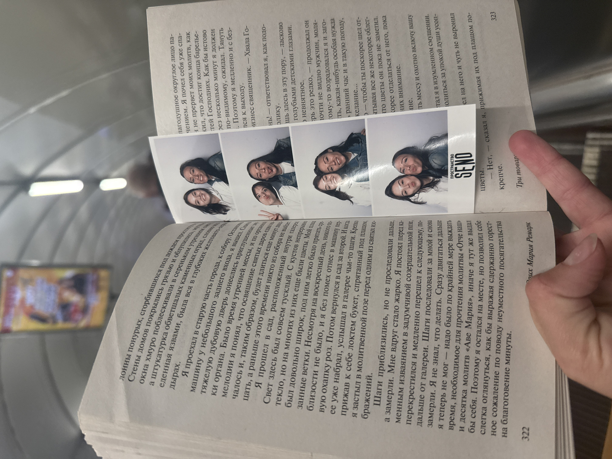 The image shows an open book lying on a surface. The book is printed in Russian. On the left side of the open book, there are several small photo booth-style pictures of two young women. The pictures are layered on top of each other and partially cover the text. The main text on the right side of the book discusses a journey and personal reflection. The last line of the text on the right page visible is "323". On the bottom left of the open book, page number "322" is visible. The text mentions "пространство SEND" which translates to "SEND space". The overall impression is a personal document, likely a diary or journal, with inserted photographs. The book is likely in a public place given the background, possibly a train station or an indoor public transit area. No specific details can confirm the location in Saint Petersburg.