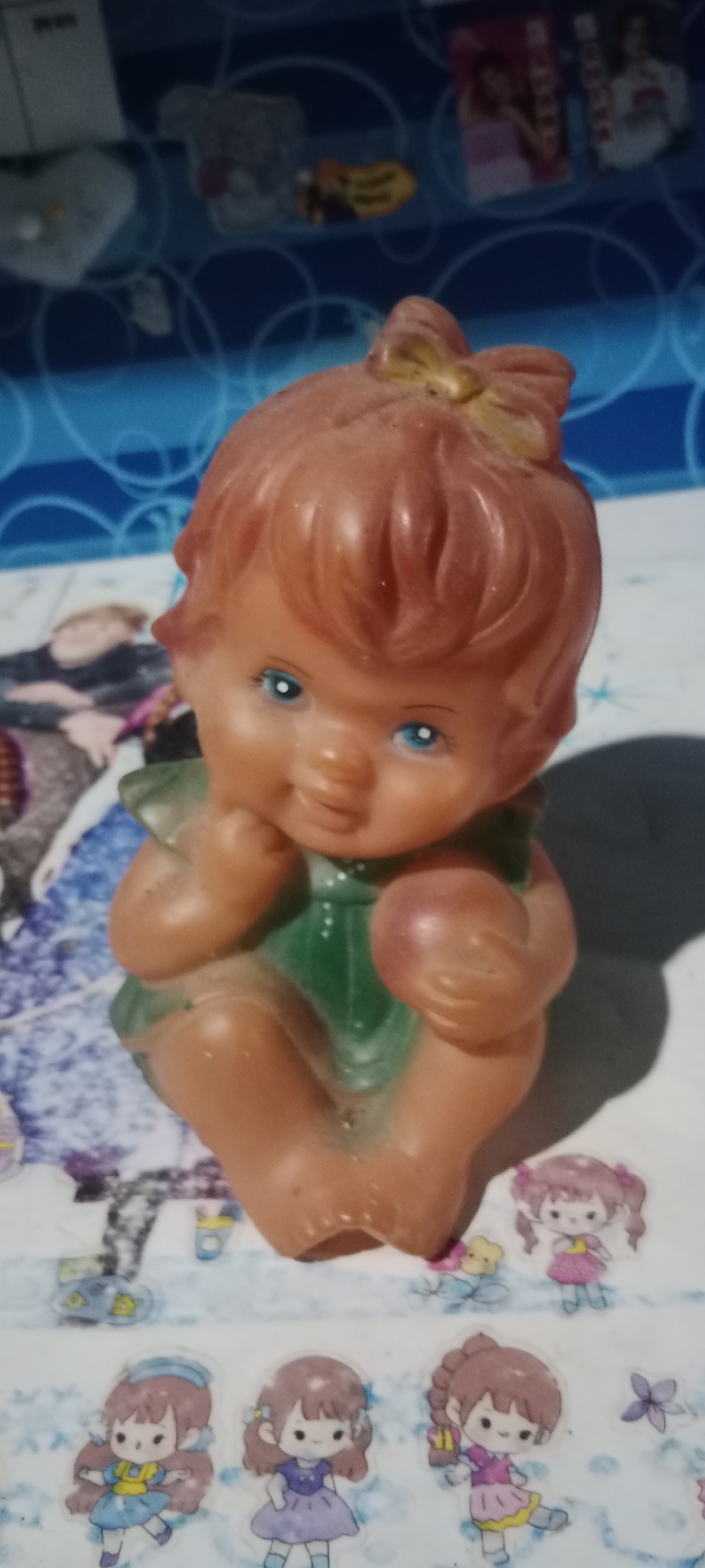 A vintage, orange-toned rubber doll with blue eyes and a yellow bow sits on a surface covered with a patterned cloth. The doll is dressed in a green outfit and is posed with its legs crossed and one arm resting on its lap. In the background, a blue patterned cloth is visible, with some blurry images of what appear to be magazines or book covers. In the foreground, below the doll, are several small stickers of cartoon-like girls in various dresses. The overall scene is indoors, with the lighting suggesting it's not a brightly lit space. There is no information in the image to suggest a specific location in the city of Pelang, Indonesia.