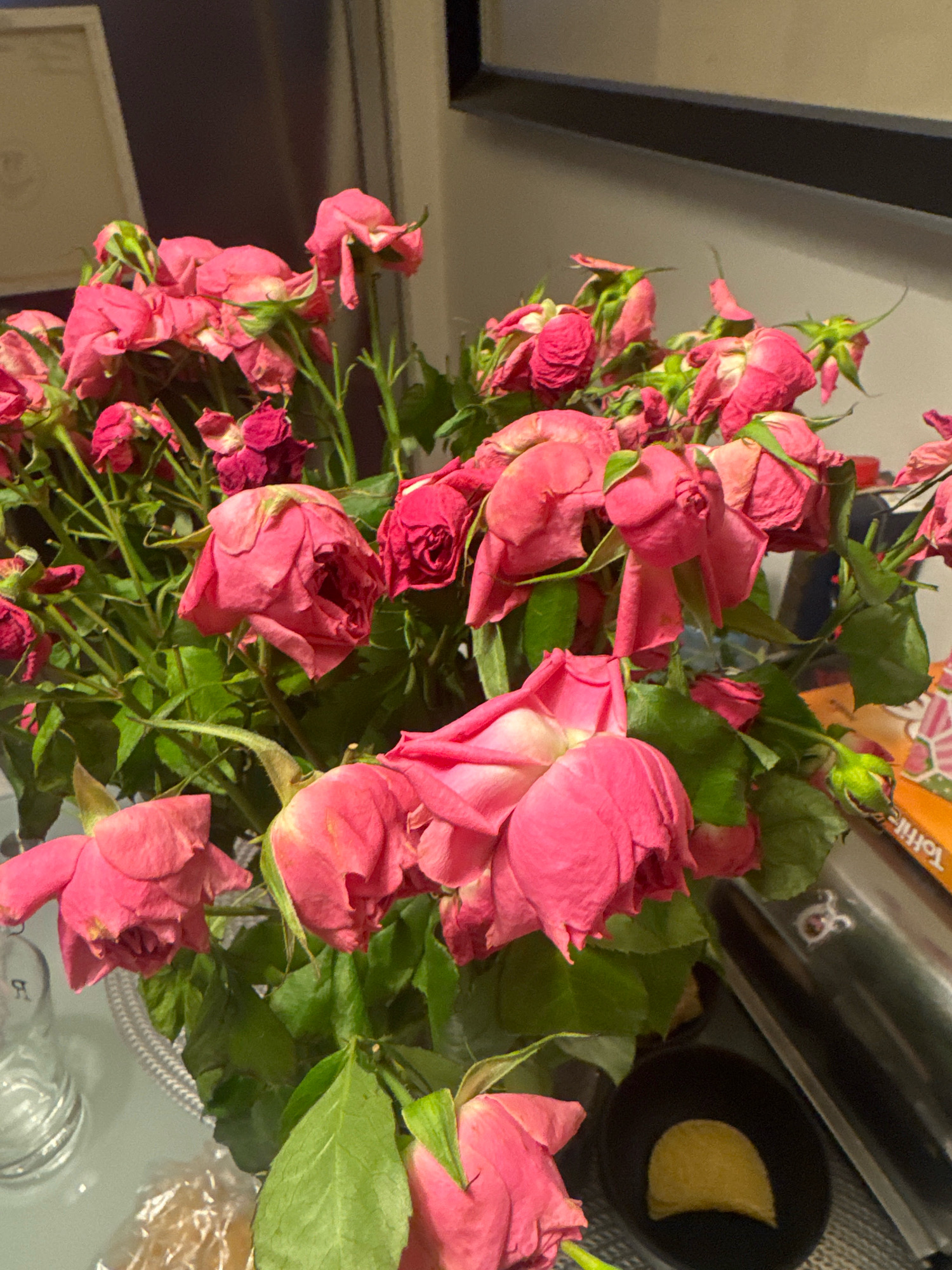 A close-up, eye-level shot captures a bouquet of pink roses, many of which appear to be wilting, with some petals curled and browned. The roses are arranged in a vase on a table. In the background, a dark, rectangular object with a lighter edge, possibly a framed picture or a shelf, is visible. To the right, a metal cylinder with text is present, and a small black dish contains a yellow, semi-circular object, possibly food. A glass object is to the left. The lighting suggests an indoor setting, possibly a home or a restaurant. There are no people visible in the image. The scene does not provide any specific indicators of its location in Almaty, Kazakhstan.