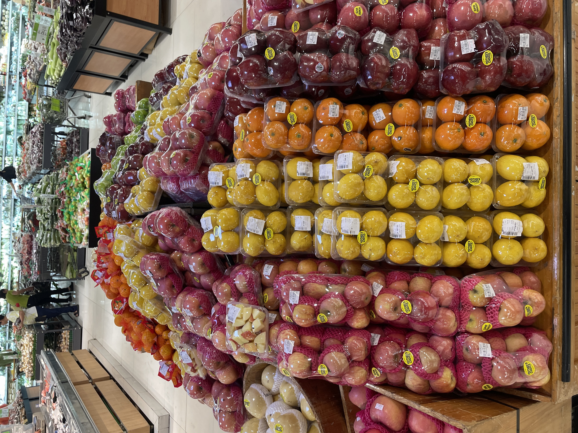 The image shows a produce section of a supermarket in Davao City, Philippines, filled with a variety of fruits displayed in plastic packaging on wooden shelves and in bins. In the foreground, there are stacks of red apples in netted bags and plastic containers. To the right, there are trays of yellow lemons and small orange fruits, possibly calamansi or kumquats. Further back, there are more apples and what appear to be pears. In the background, other shoppers can be seen browsing the aisles, and shelves stocked with vegetables like eggplants and gourds are visible. The overall scene is one of a well-stocked grocery store.