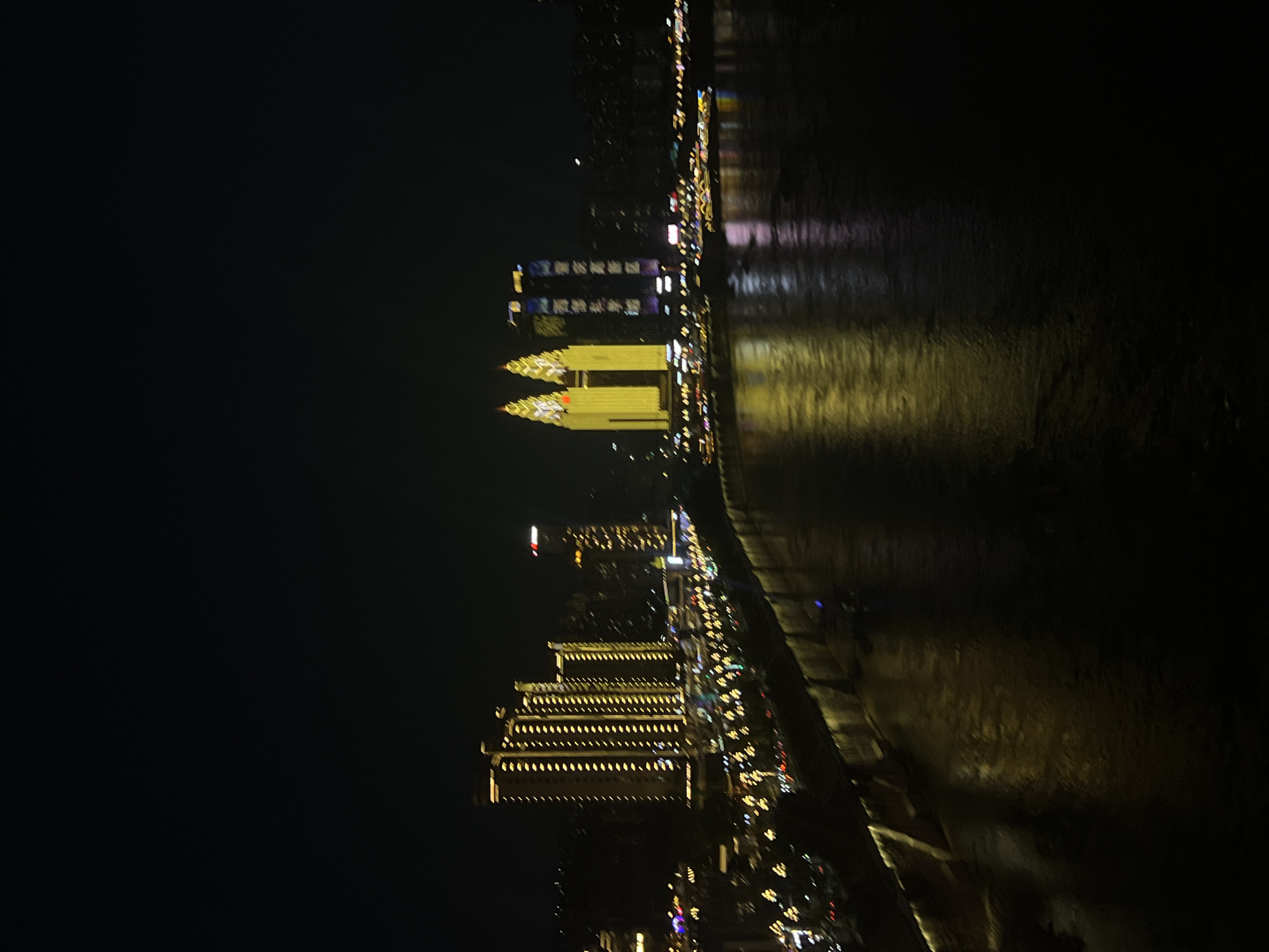 This is a nighttime long-exposure photograph of a city skyline at the Nan'an District, Chongqing, China, specifically the area along the Yangtze River. The image captures a waterfront promenade lined with buildings illuminated by yellow and white lights. In the middle ground, a cluster of tall, rectangular buildings with a grid pattern of lights is prominent. To the right, two distinctive twin towers are brightly lit in yellow, resembling pointed hats. Further in the background, other buildings create a denser urban silhouette. The river in the foreground reflects the city lights, creating shimmering, elongated streaks of yellow and white on its dark surface. A bridge or elevated walkway runs along the waterfront, also illuminated. The overall impression is one of a vibrant, modern cityscape at night, with the water serving as a dynamic canvas for the reflected urban glow. There are no visible people or vehicles in the immediate foreground or identifiable subjects within the frame.