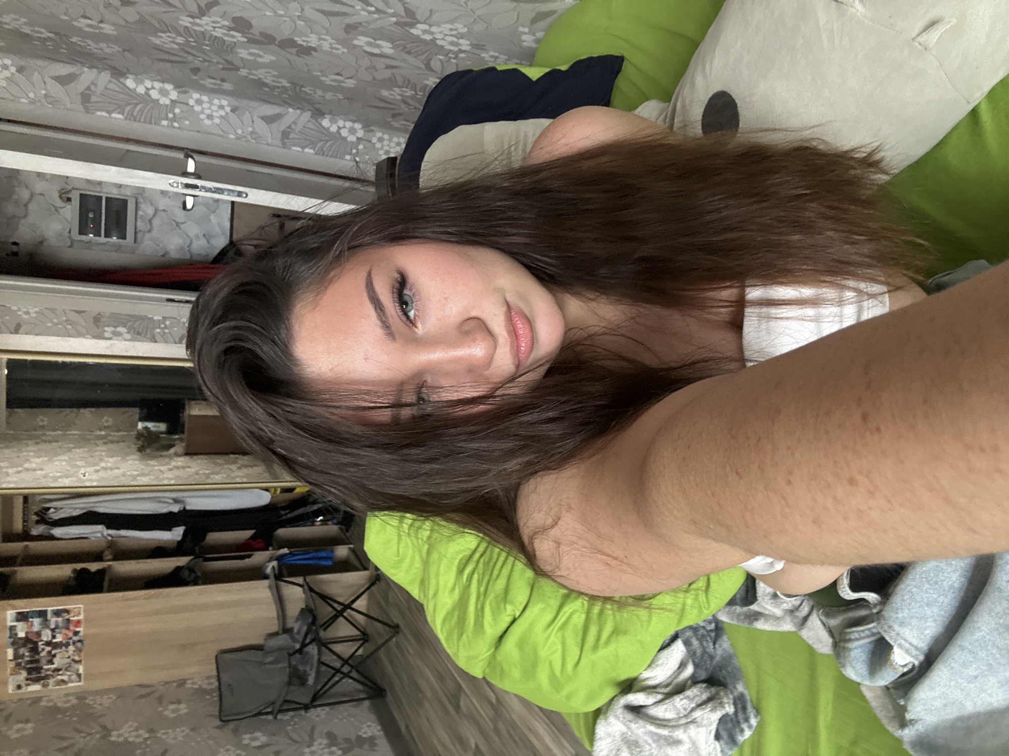 A young woman with long, dark brown hair is taking a selfie while lying on a green bed. She is wearing a white top. Her left arm is extended towards the camera, and her face is turned to the right. The background shows a bedroom with a closet, a chair, and patterned wallpaper. There is no visible indication of the city of Perm, Russia.