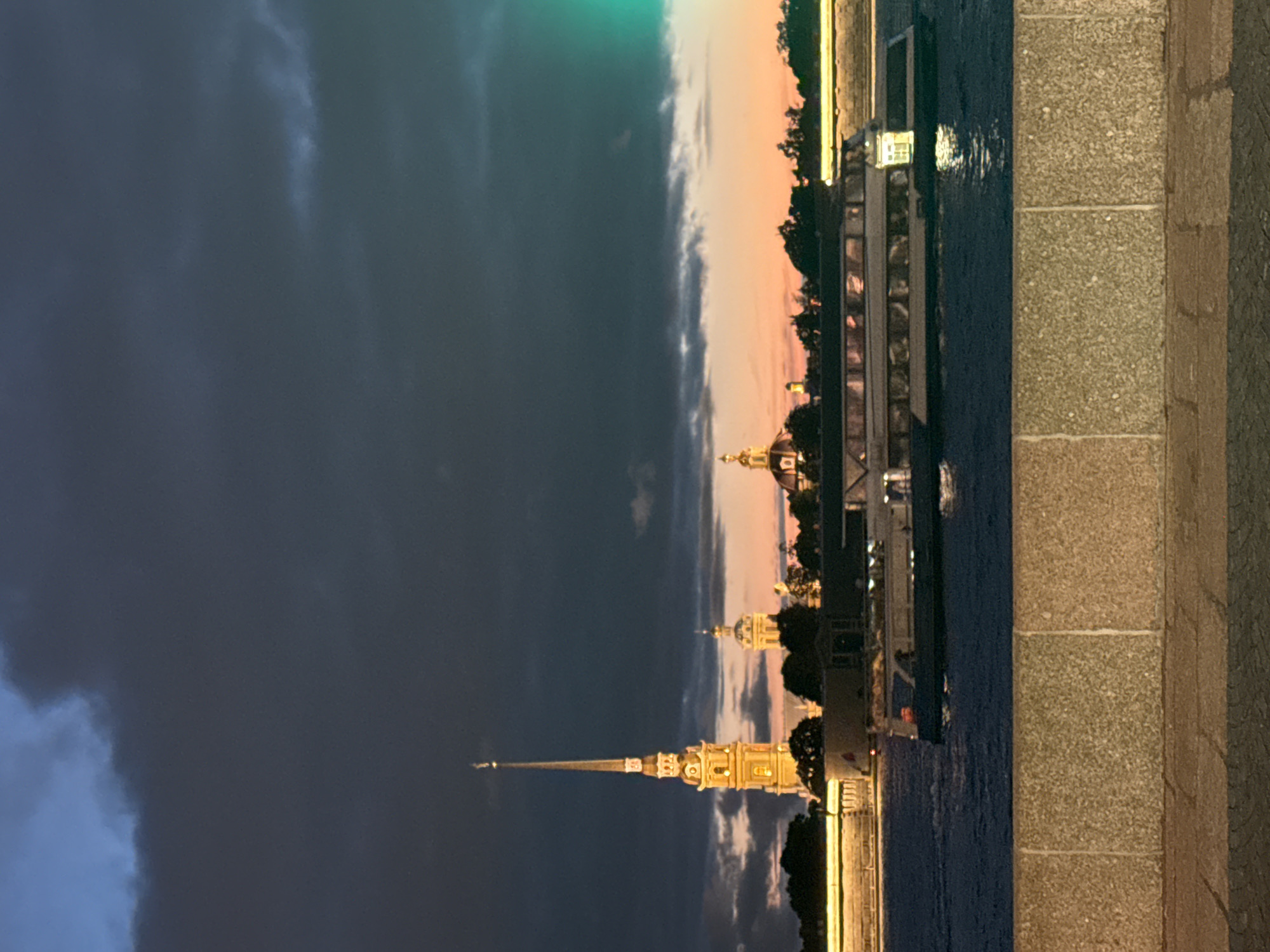 An evening shot shows a river with a large boat and a skyline of buildings. The sky is dark with clouds, and the setting sun casts a warm glow behind the buildings. A tall, illuminated spire with a pointed top stands out among the structures. The boat, which appears to be a tour boat, is lit up from the inside and is moving on the water. The foreground shows a stone embankment and pavement.