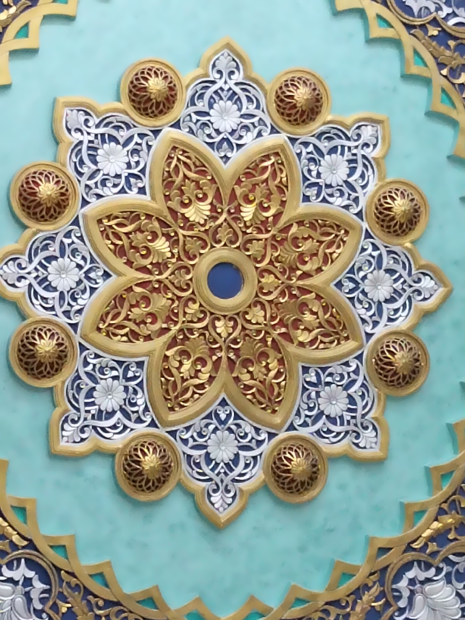 The image shows a close-up of an ornate architectural detail, likely a ceiling or wall decoration, in Kagan, Uzbekistan. The design features a central, eight-petaled floral motif with intricate gold filigree against a deep red background. This central element is surrounded by layers of relief carvings in white and silver, depicting smaller floral patterns and scrolling foliage. Scattered around the main floral design are eight circular decorative elements, each with a gold, openwork lattice pattern over a red interior. The entire design is set against a textured turquoise background, with additional geometric gold and dark blue bordering visible at the edges of the frame, suggesting a larger, elaborate decorative scheme. The lighting appears to be natural, highlighting the three-dimensional aspects of the carvings. No people are present in the image.